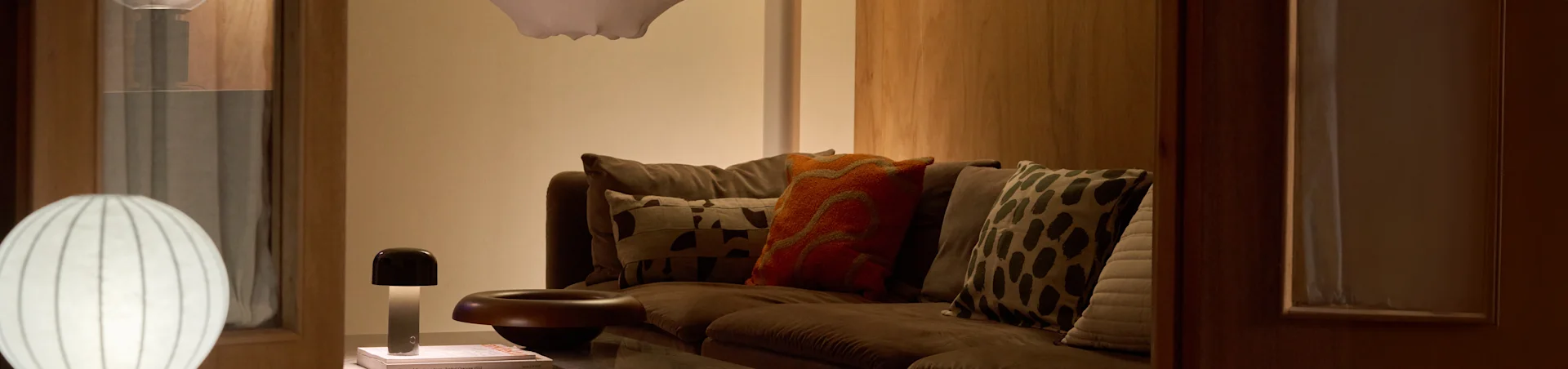 Ein gemütliches Wohnzimmer mit einem braunen Sofa, verschiedenen gemusterten Kissen und zwei Lampen: einer großen weißen gerippten Stehlampe und einer kleinen pilzförmigen Tischlampe.