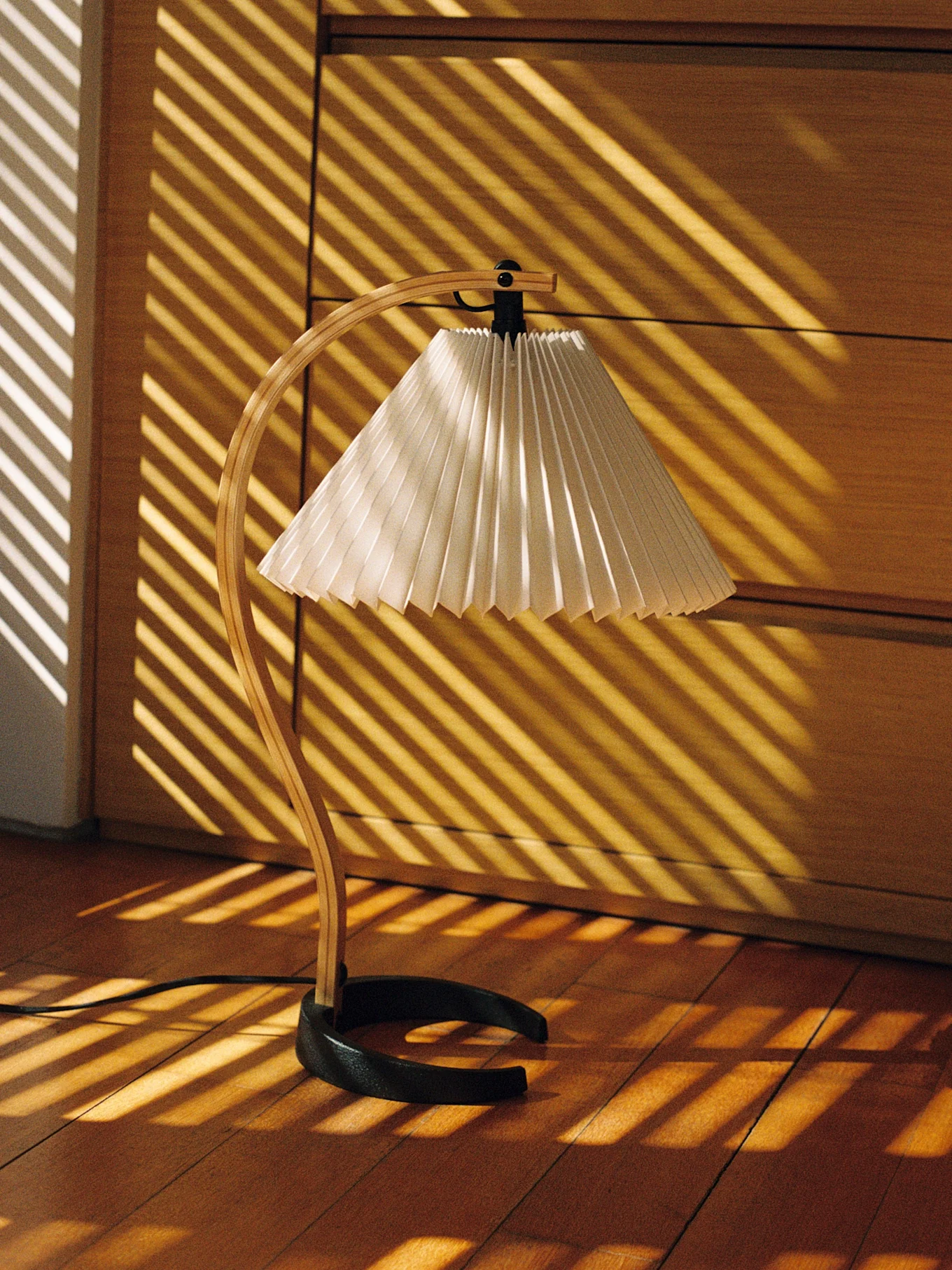 Schreibtischlampe mit gebogenem Holzständer und plissiertem Schirm, beleuchtet von diagonalen Sonnenlichtstreifen auf Holzfußboden.