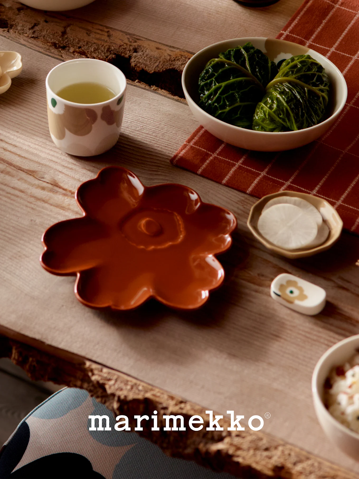 Tisch gedeckt mit Marimekko-Geschirr: ein rostfarbener Blütenteller, eine Tasse Grüntee, Kohlrouladen und geschnittene Radieschen.