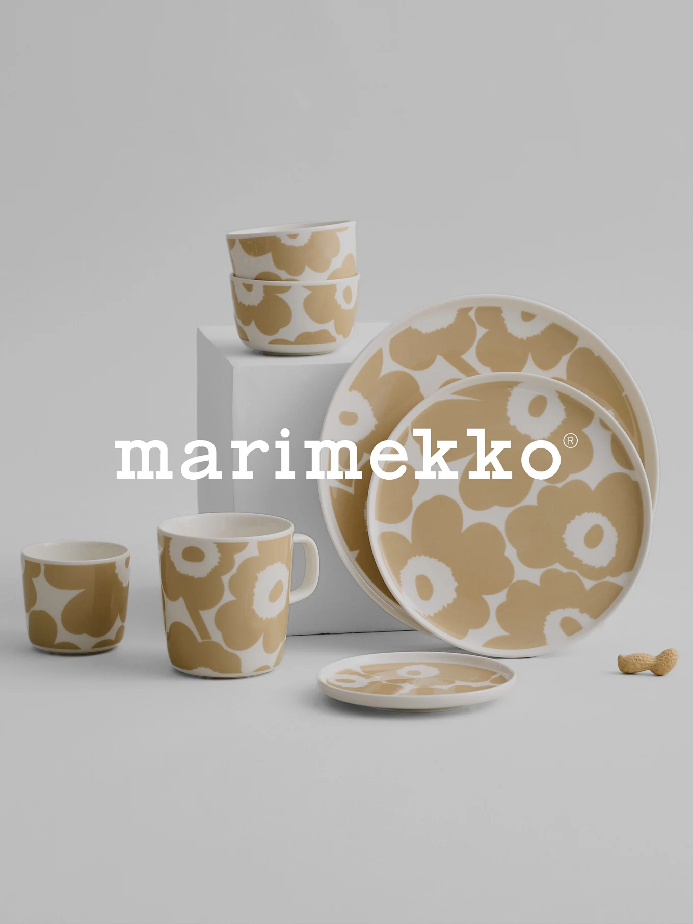 Marimekko Geschirrset mit beigem und weißem Blumenmuster, bestehend aus Schüsseln, Tasse, Bechern, Tellern und einer Erdnuss auf grauem Untergrund.