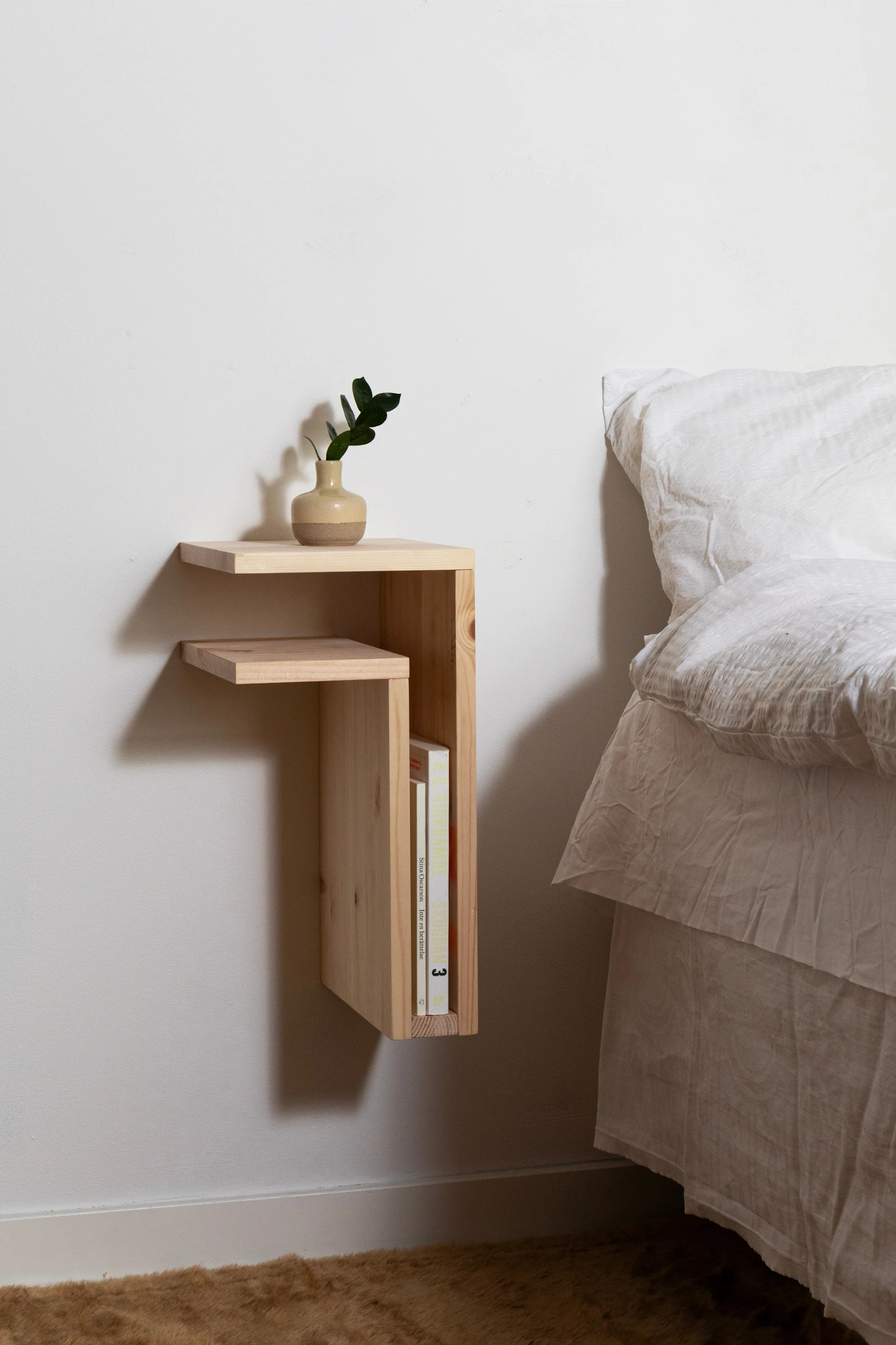 Ein schwebender Nachttisch aus hellem Holz mit einer Pflanze in einer zweifarbigen Vase und Büchern, neben einem Bett mit weißer Bettwäsche.