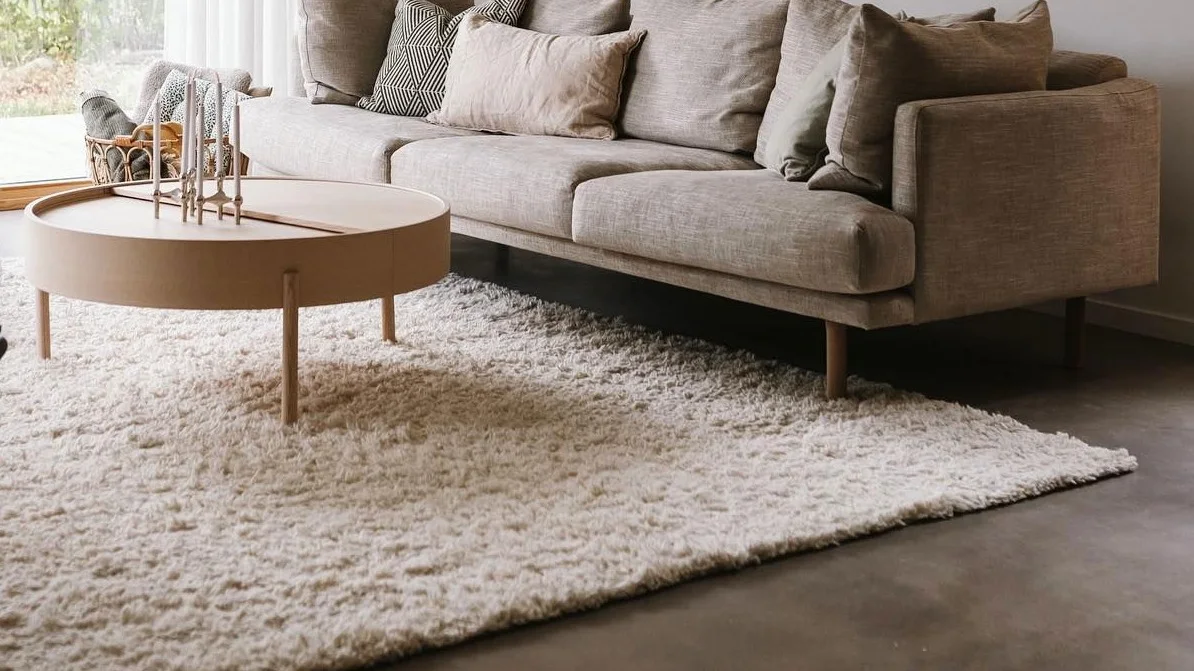 Ein Wohnzimmer in beige mit dem Sjövik Sofa von 1898, davor steht der Arc Couchtisch von Woud und darunter liegt der helle Cozy Wollteppich von Scandi Living.