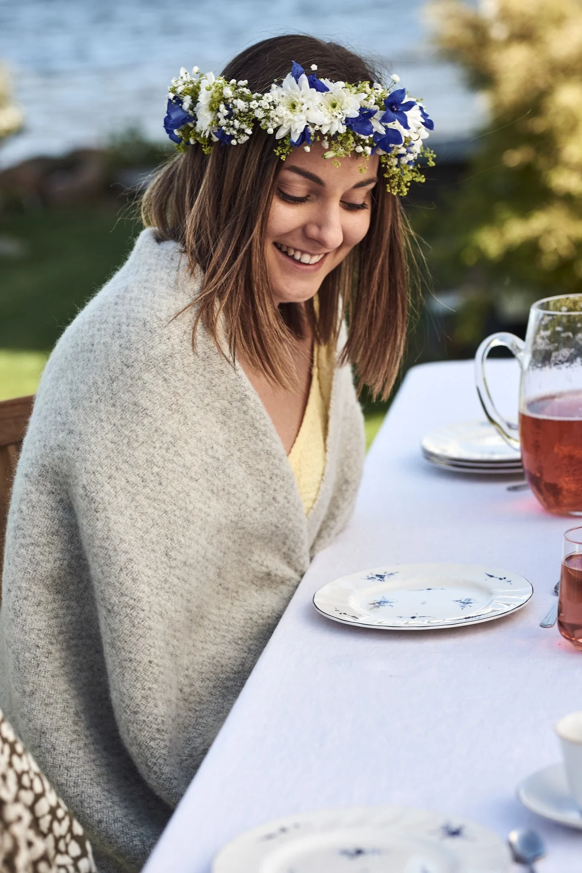 Eine Frau mit Blumenkranz sitzt draußen an einem gedeckten Tisch und feiert Midsommar in Schweden.