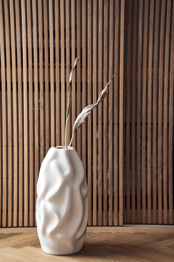 Dunkle Hölzer und gerade Linien, die auf runde Formen treffen sind Teil der Wohntrends fur den Herbst 2021. Hier steht die schöne Drift Vase von Cooee Design in Beige vor einer dunklen Holzwand.