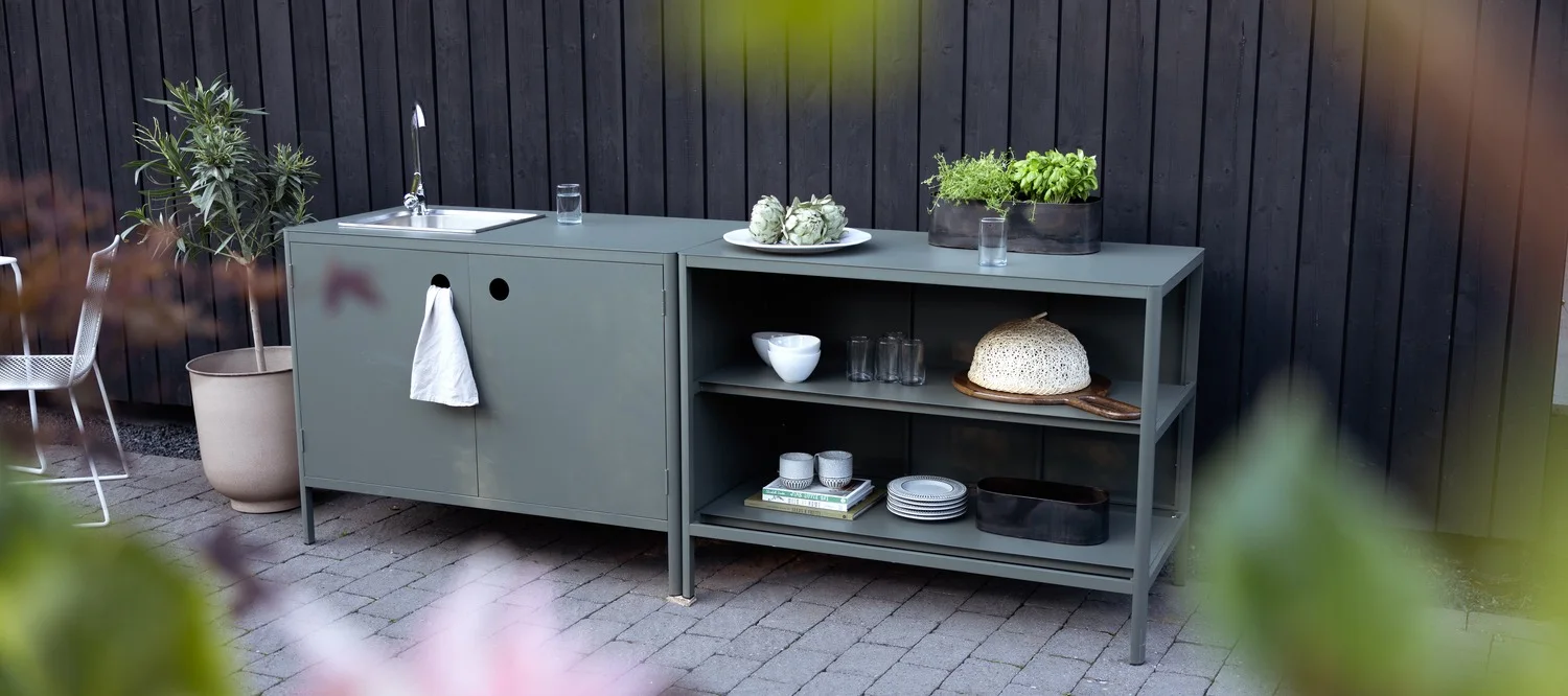 Eine graugrüne Outdoor-Küchenzeile mit Spüle und Handtuch links, sowie Regalen mit Geschirr und Topfkräutern rechts.