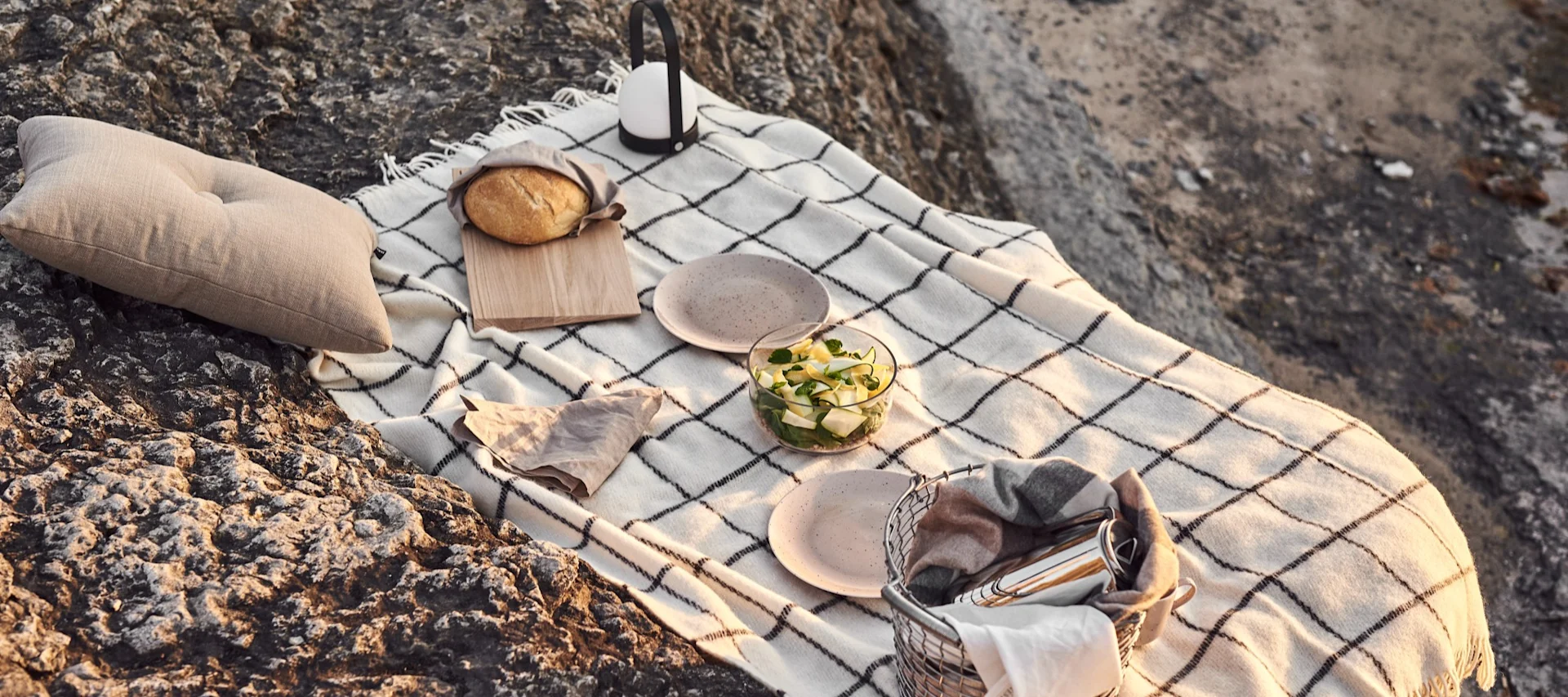 Gemütliches Picknick auf Felsen mit karierter Decke, beige Kissen, Brot, Salat, Tellern, Laterne und Picknickkorb.