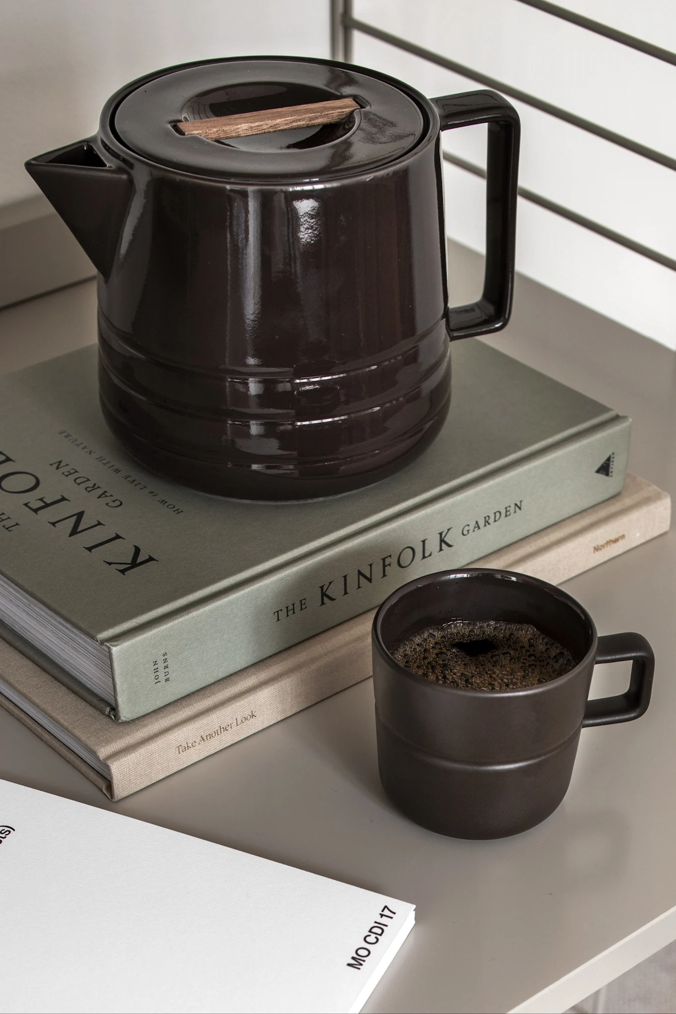 Dunkelbraune Keramik-Kaffeekanne mit Holzakzent auf dem Deckel, passender Becher mit Kaffee und zwei Büchern.