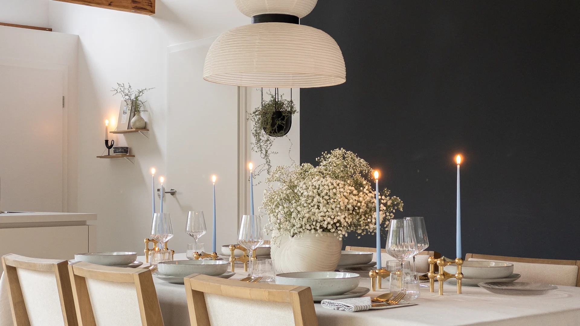 Eleganter Esstisch mit weißer Tischdecke, grauen Gedecken, goldenem Besteck, brennenden blauen Kerzen und weißen Schleierkrautblumen.