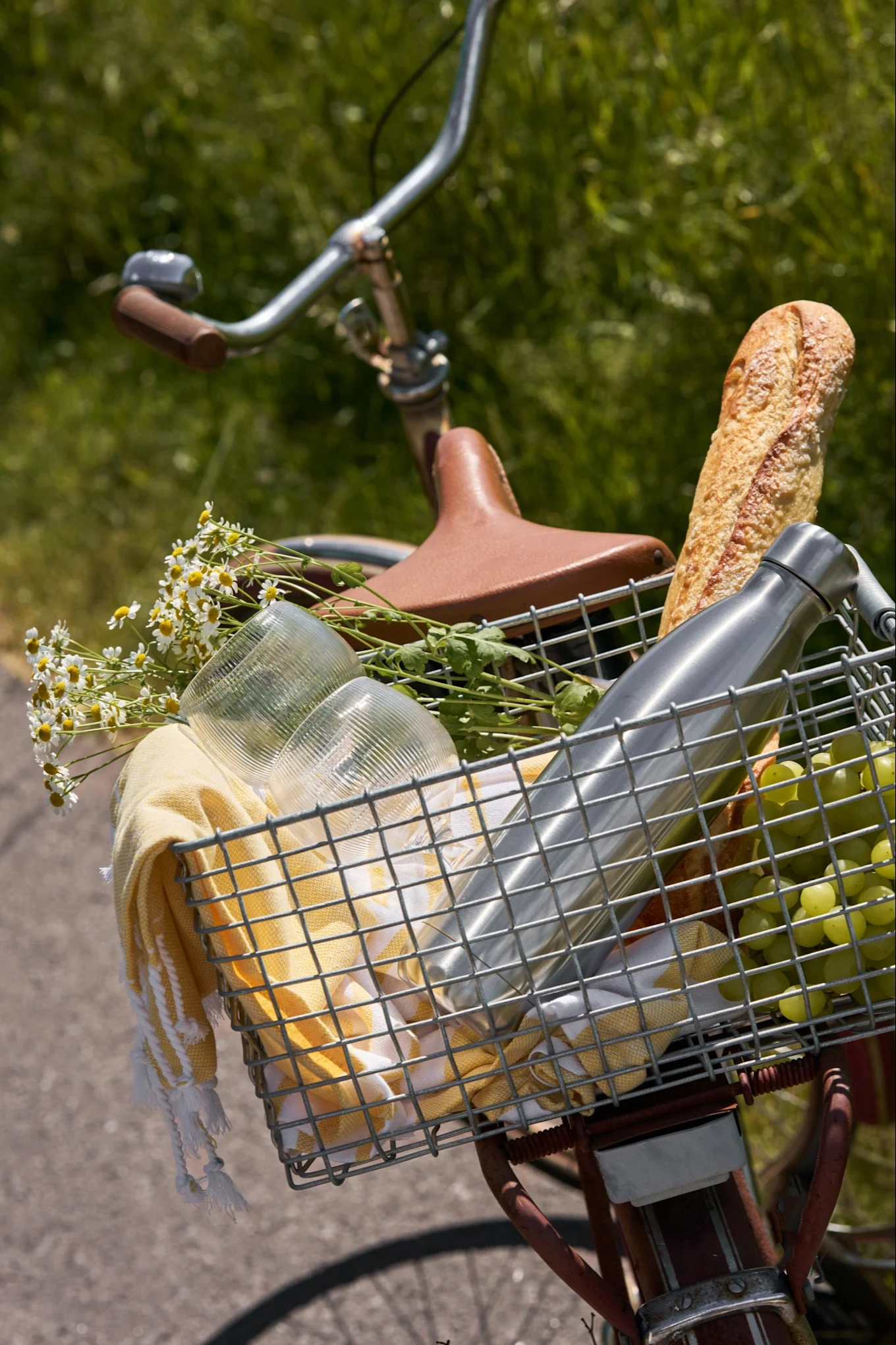 Ein Fahrradkorb mit Picknickartikeln: Baguette, Wasserflasche, Trauben, Becher, gelbes Handtuch und weiße Gänseblümchen.