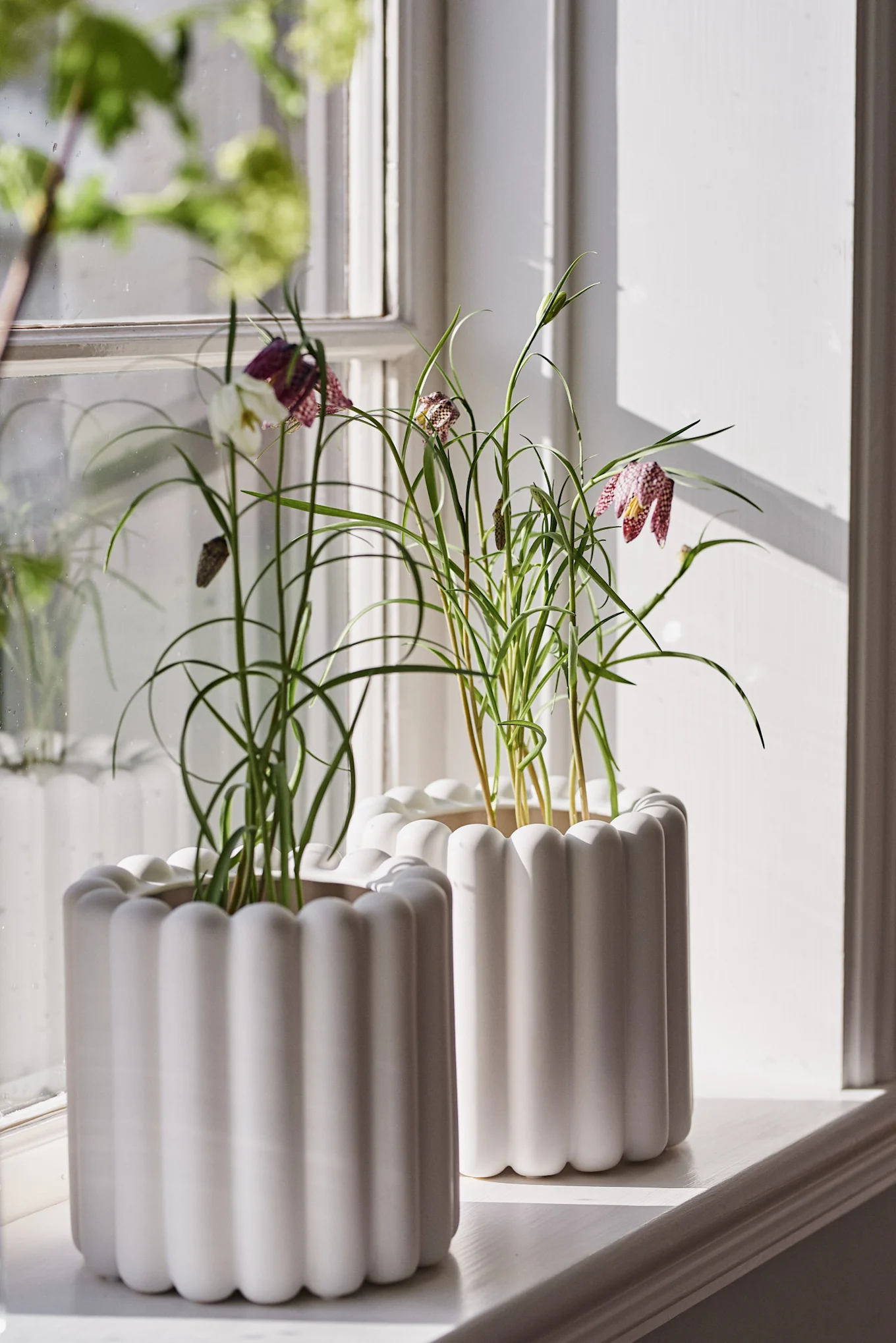 Zwei weiße Blumentöpfe aus der Mist-Kollektion von DBKD auf einer Fensterbank, bestückt mit frühlingshaften Blumen.