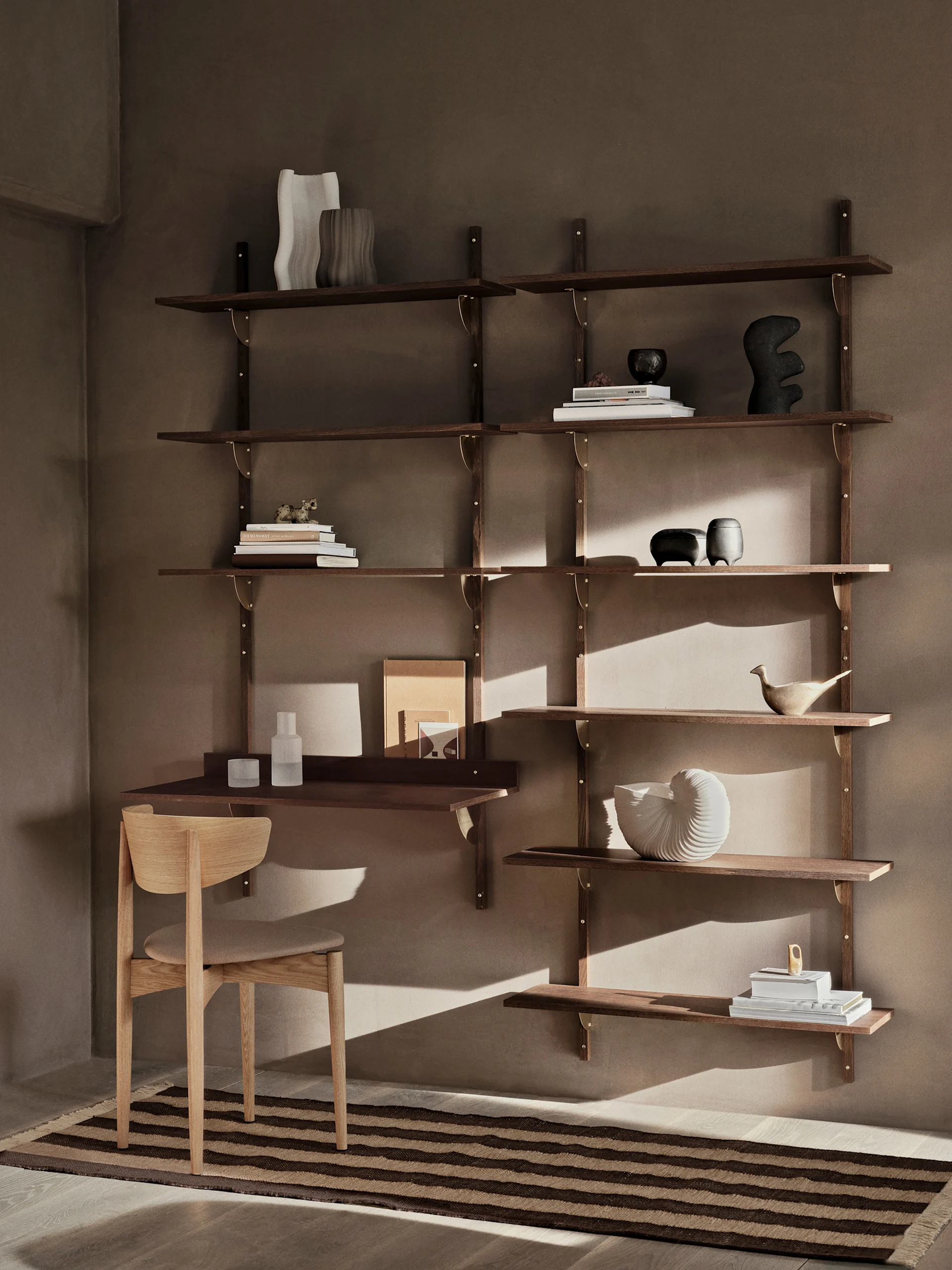 Ein modernes Wandregal aus dunklem Holz mit Schreibtisch und Stuhl, verziert mit Büchern und Deko-Objekten, steht vor einer warmbraunen Wand mit einem gestreiften Teppich.