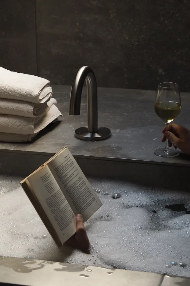 Hand hält offenes Buch in einem Schaumbad, mit Weinglas, weißen Handtüchern und einem modernen silbernen Wasserhahn auf dem Sims.