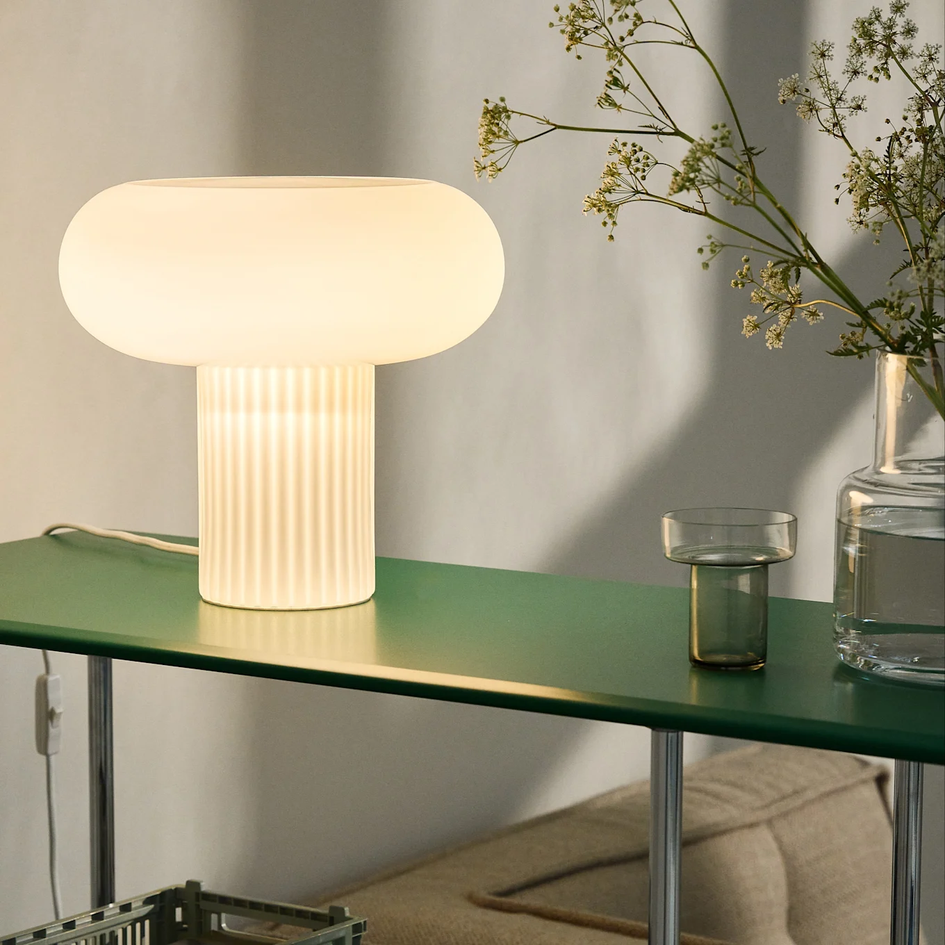 Leuchtende pilzförmige Tischlampe mit geripptem Sockel auf grünem Regal, daneben Blumen in einer Vase.
