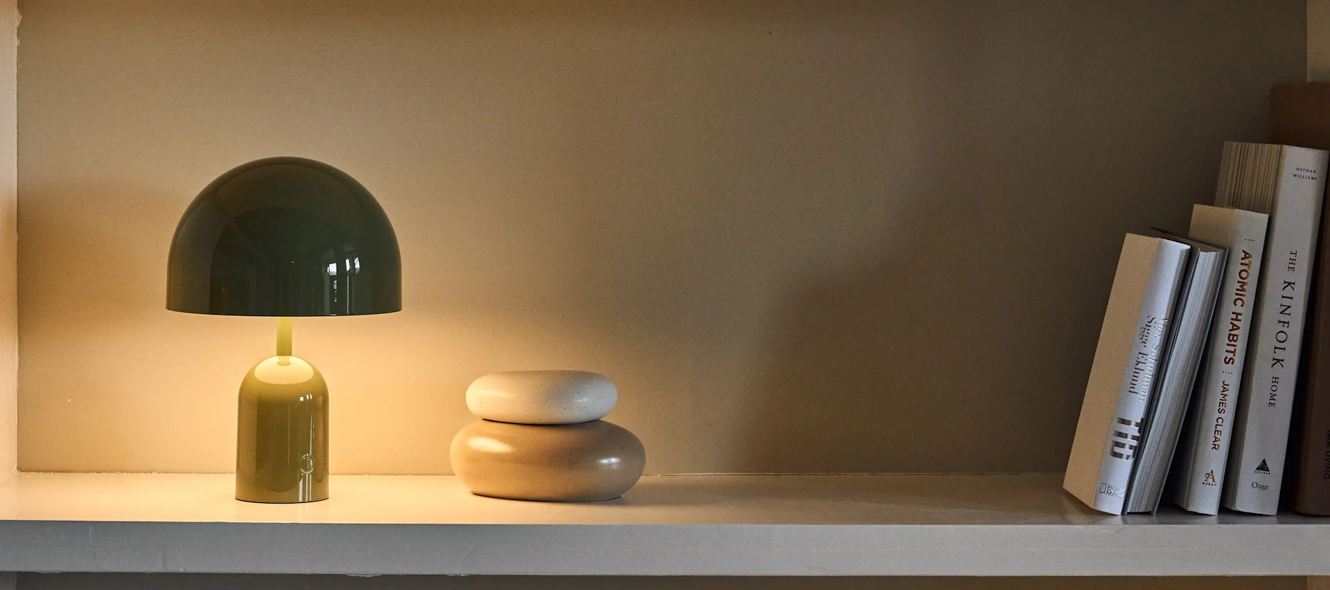 Grüne, pilzförmige Tischlampe mit warmem Licht auf einem Regal, daneben zwei gestapelte ovale Ziersteine und eine Reihe heller Bücher.