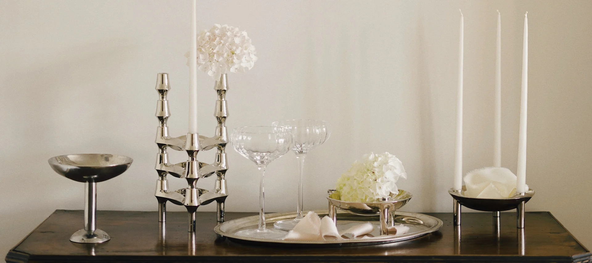 Elegante Tischdekoration mit silbernen Kerzenhaltern, weißen Kerzen, Gläsern und weißen Hortensien auf Holzoberfläche.