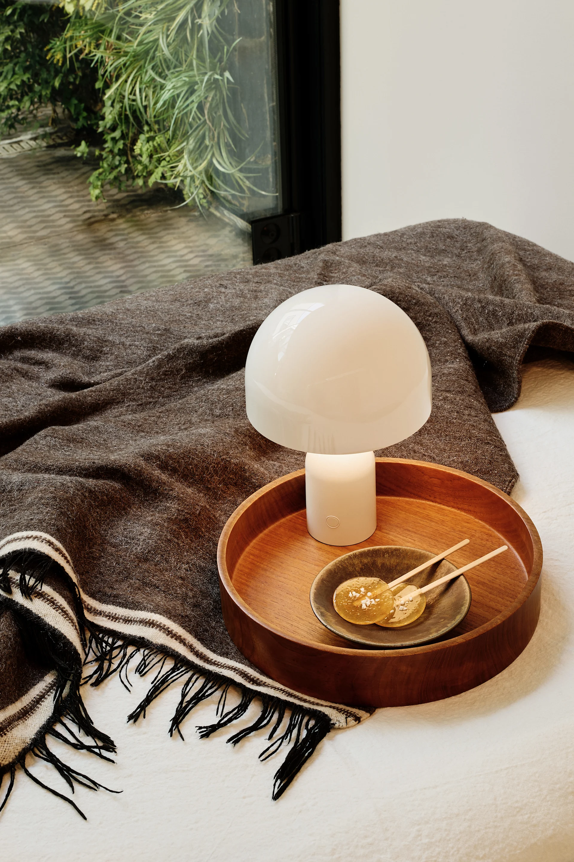 Weiße Akku-Tischleuchte auf einer dunkelbraunen Decke mit einem Holztablett, das zwei Lollies enthält.