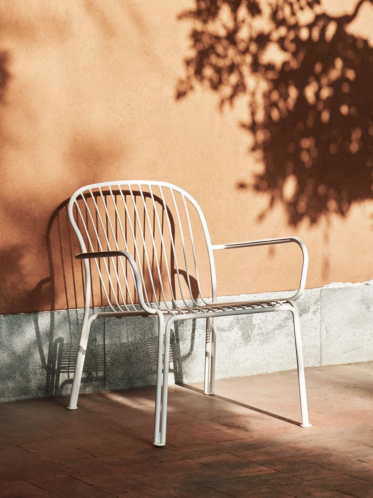 Weißer Gartenstuhl aus Metall mit Lamellenlehne vor einer orangen Wand mit Baumschatten an einem sonnigen Tag.