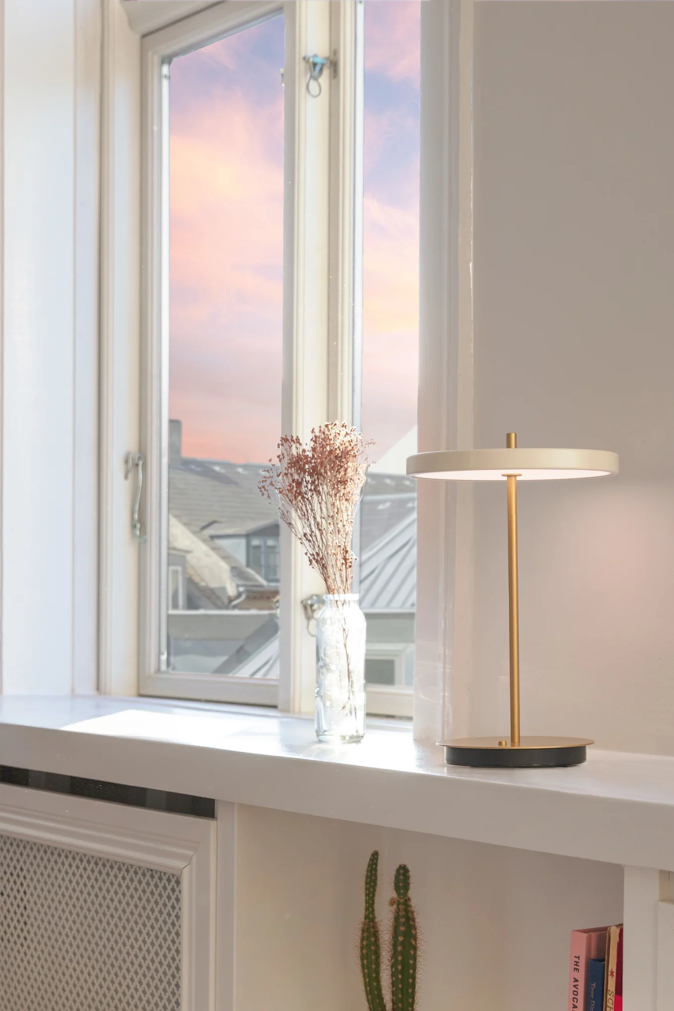 Moderne Tischlampe mit goldenem Stiel neben Trockenblumen auf einem weißen Regal vor einem weichen Sonnenuntergangshimmel.