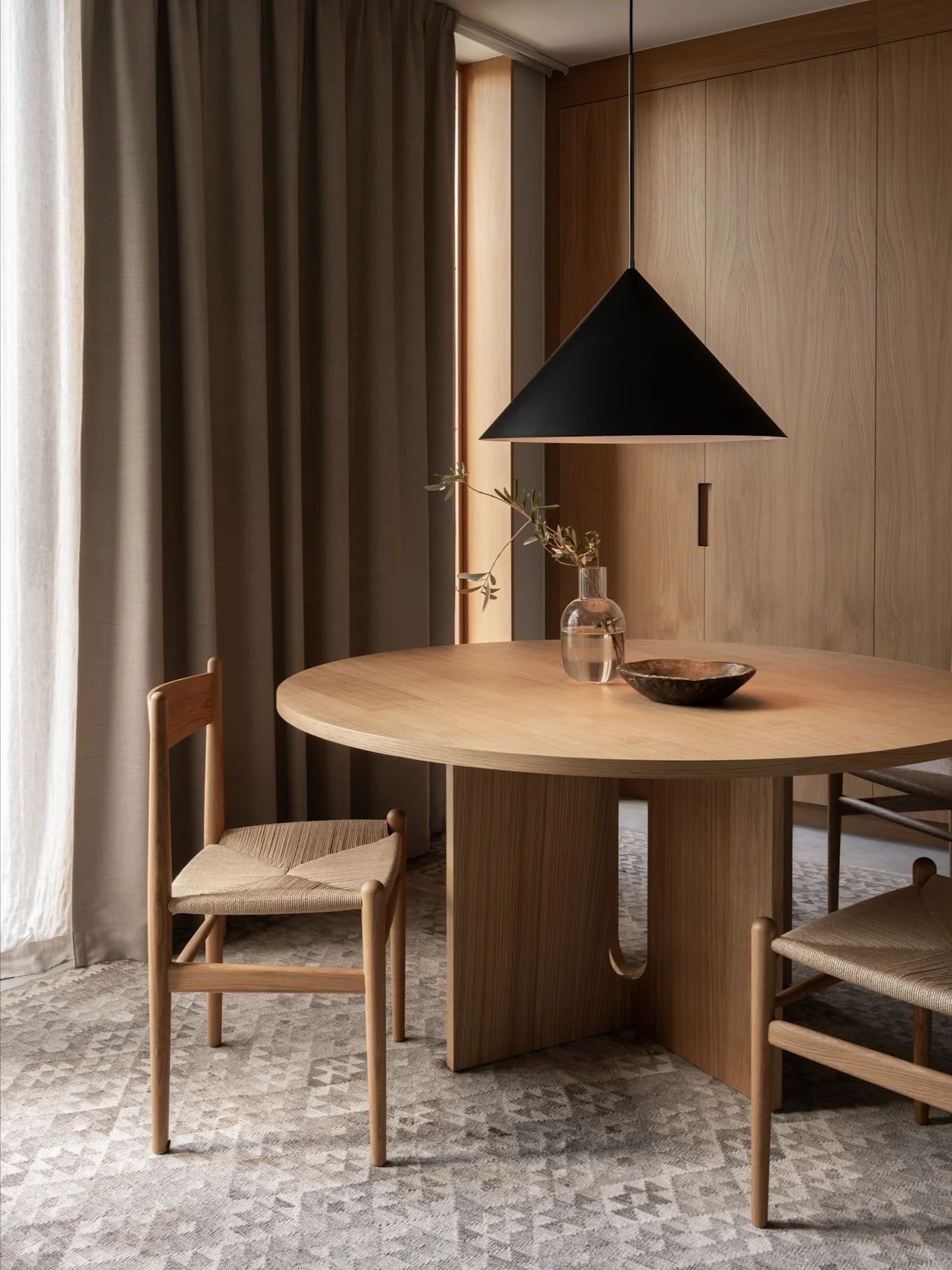 Ein minimalistischer Essbereich mit rundem Holztisch, zwei Stühlen mit geflochtenen Sitzen, schwarzer Pendelleuchte und Holzwänden.