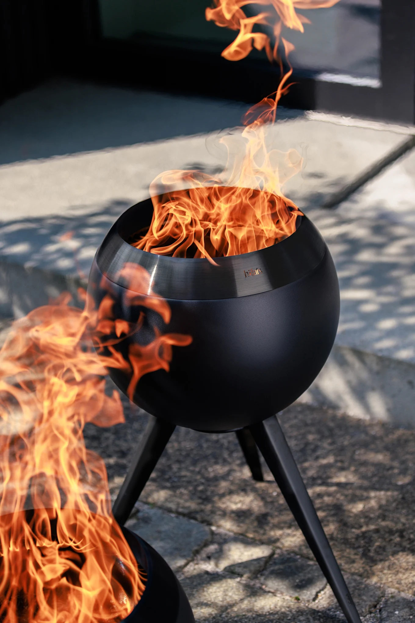 Zwei schwarze, kugelförmige Outdoor-Feuerstellen mit hellen orangefarbenen Flammen auf einer gepflasterten Terrasse.