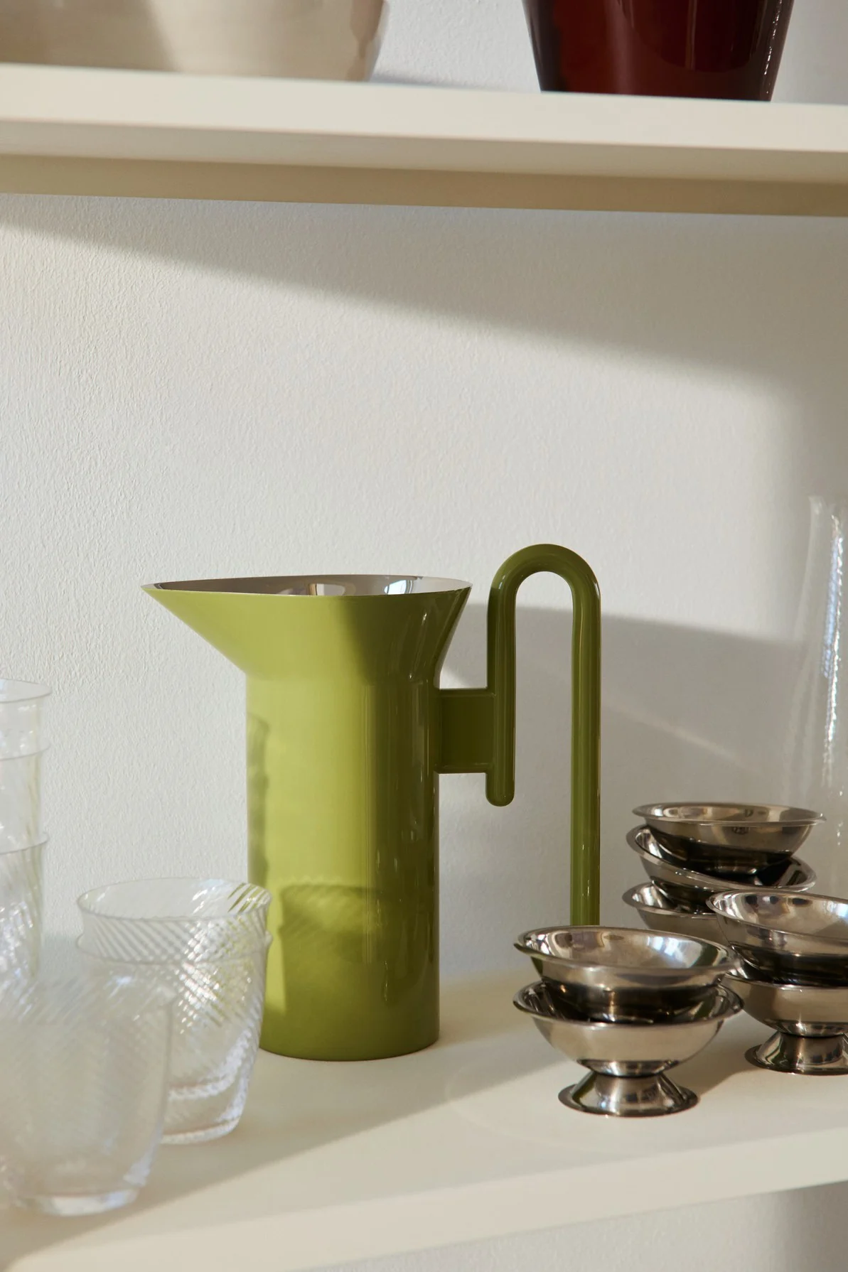 Olivgrüne Kanne mit U-förmigem Griff, flankiert von klaren gemusterten Gläsern und Stapeln kleiner silberner Dessertschalen auf einem Regal.