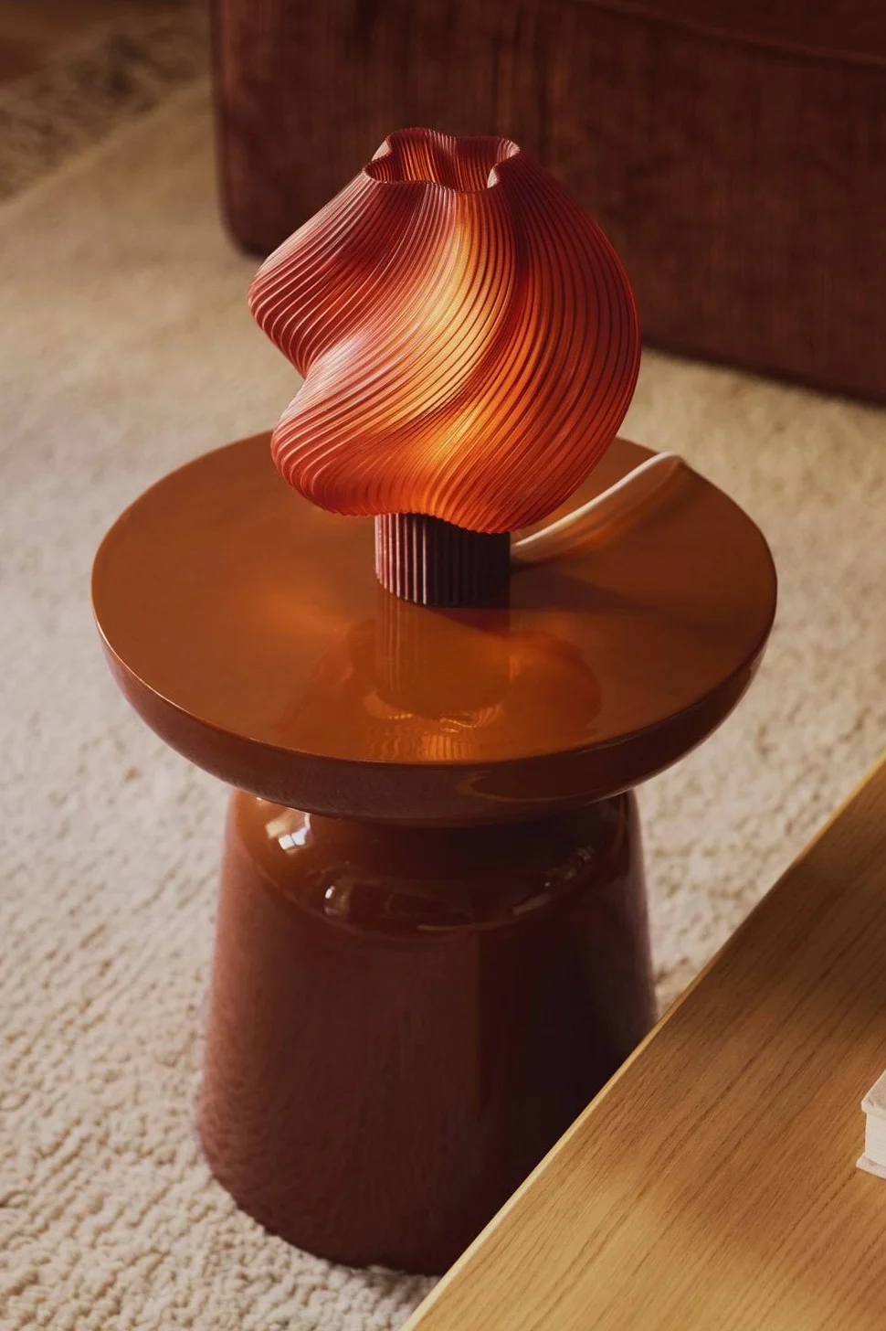 Eine warm beleuchtete, wellige orangefarbene Plisseelampe steht auf einem glänzenden braunen Beistelltisch.