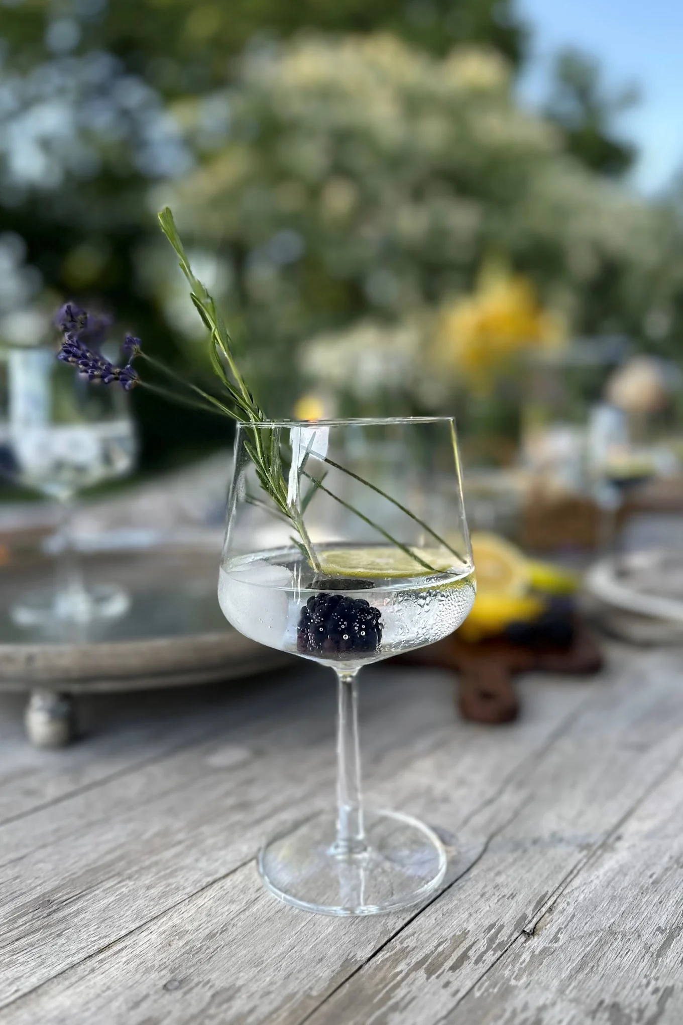 Ein klares Stielglas auf einem Holztisch, das eine Brombeere, eine Limettenscheibe, einen Lavendelzweig und klare Flüssigkeit enthält.