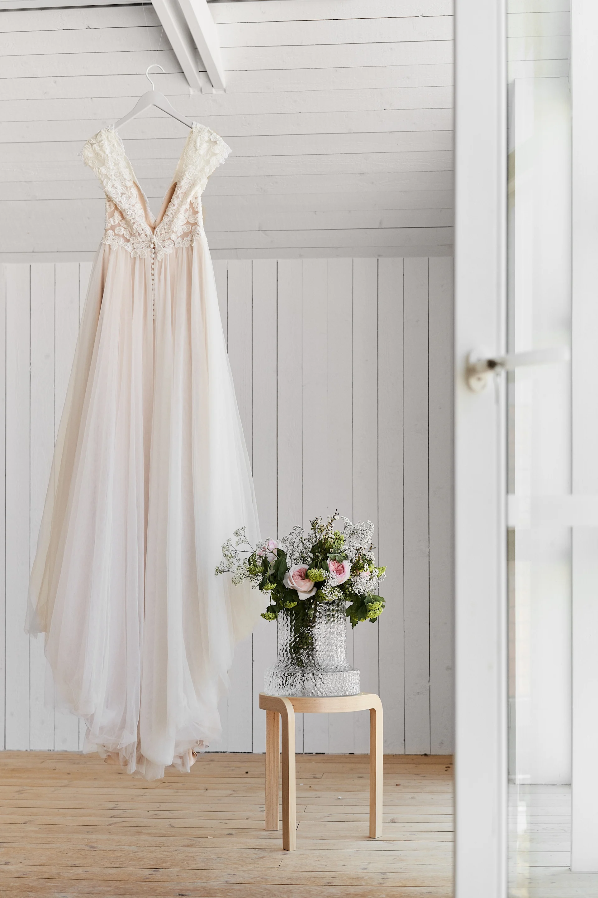Einzigartige Hochzeitsideen müssen fotografiert werden, um in Erinnerung zu bleiben. Füllen Sie Vasen mit schönen Blumen, um Ihre Fotos aufzuwerten. Hier sehen Sie ein Hochzeitskleid, das neben einem Holzhocker und einer Vase hängt.
