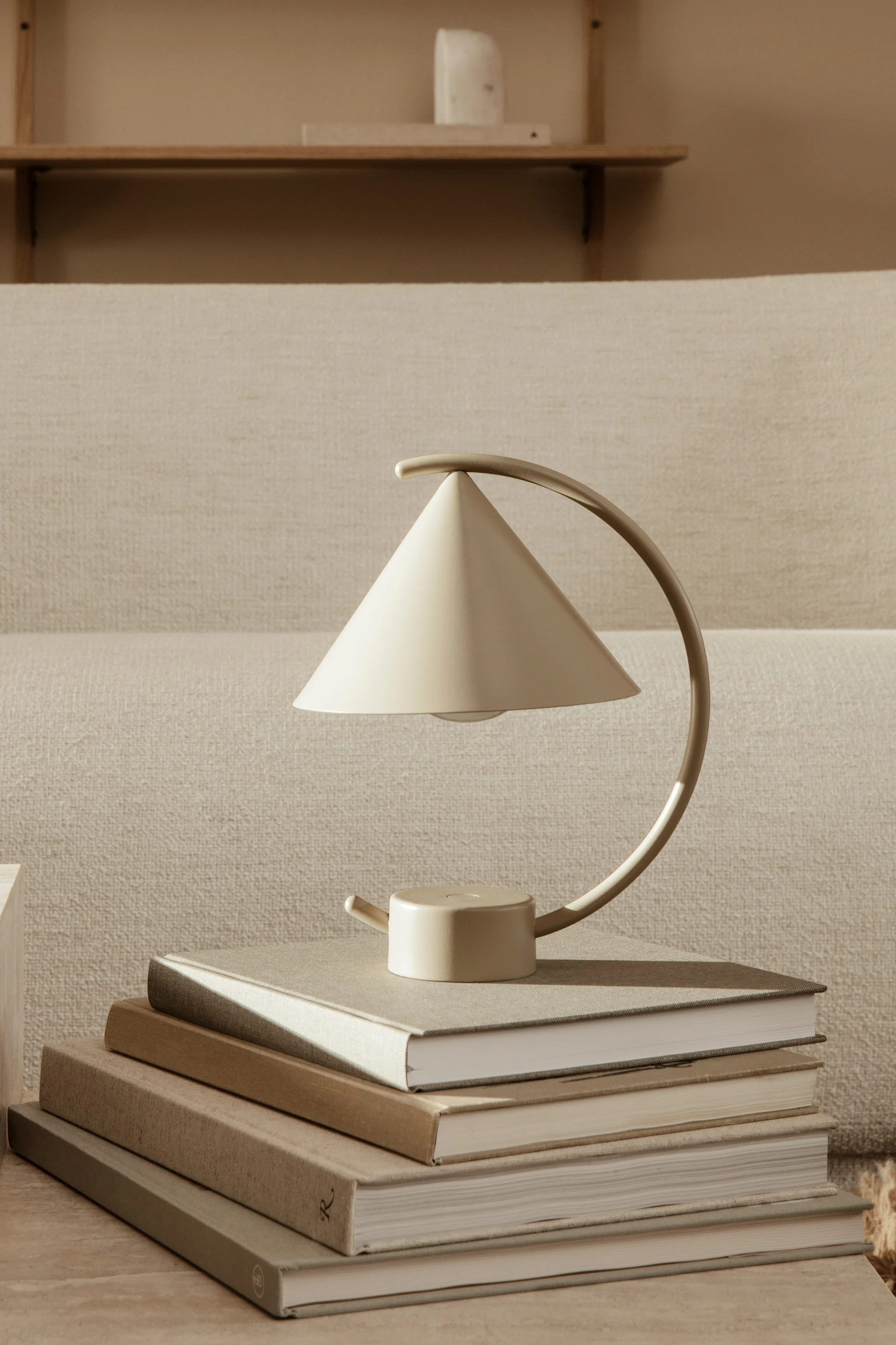 Eine beige konische Akku-Tischleuchte mit gebogenem Griff steht auf einem Stapel neutralfarbener Bücher in minimalistischer Umgebung.