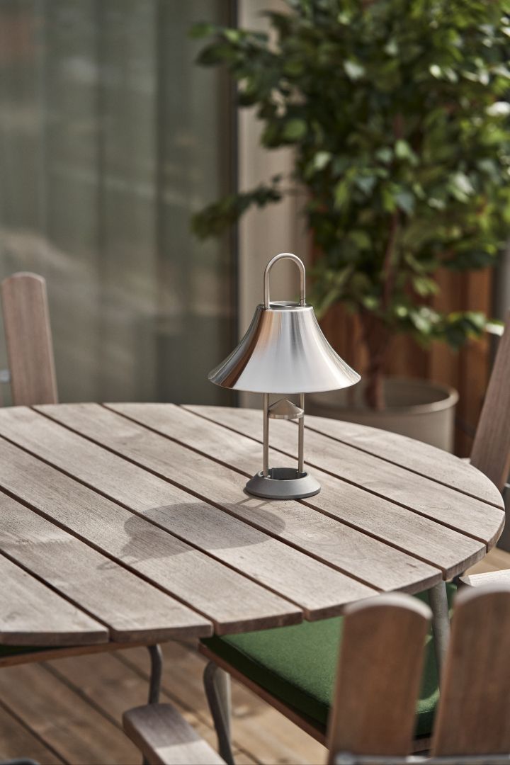 Bringen Sie stimmungsvolles Licht in Ihren Garten oder auf Ihre Terrasse mit der tragbaren Lampe Mousqueton aus Edelstahl von HAY, die auf einem runden Teakholz-Außentisch steht.