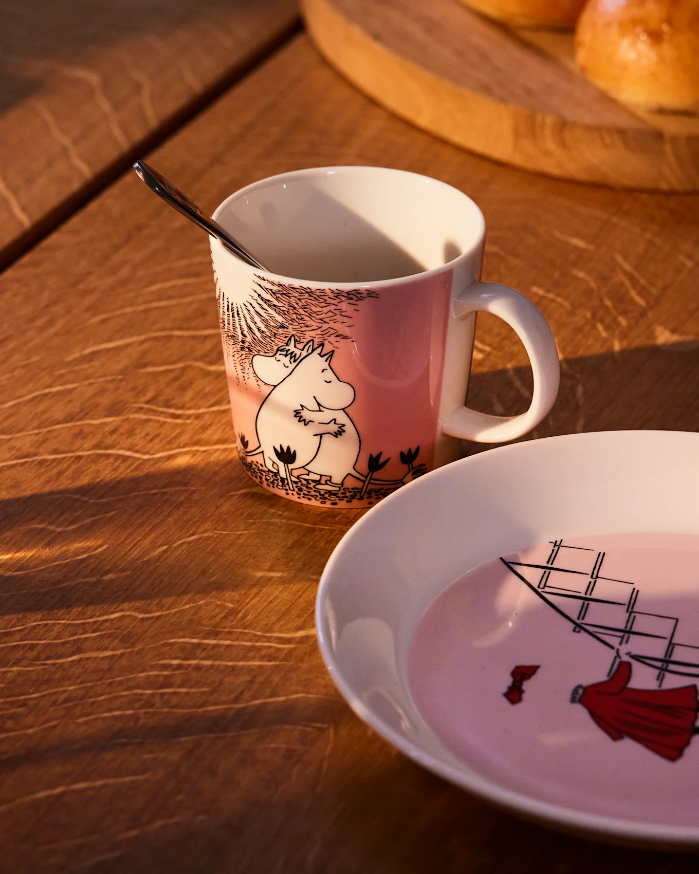 Rosa Moomin-Tasse mit Moomintroll und Snorkfräulein, daneben ein passender Teller mit Segelschiff, auf einem Holztisch.