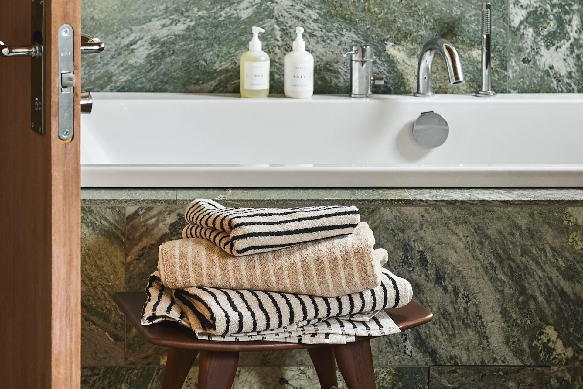 Ein Stapel beige und gestreifte Handtücher auf einem Holzschemel in einem Badezimmer mit einer weißen Badewanne und grünen Marmorwänden.