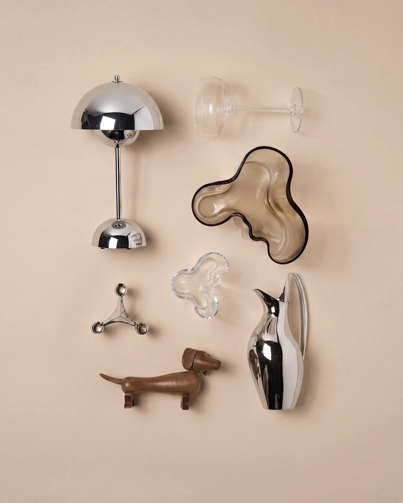 Moderne Wohndekoration: Chrom-Pilzlampe, klares Weinglas, braune Wellenschale, silberner Kerzenhalter, Dackel aus Holz, Silberkanne.