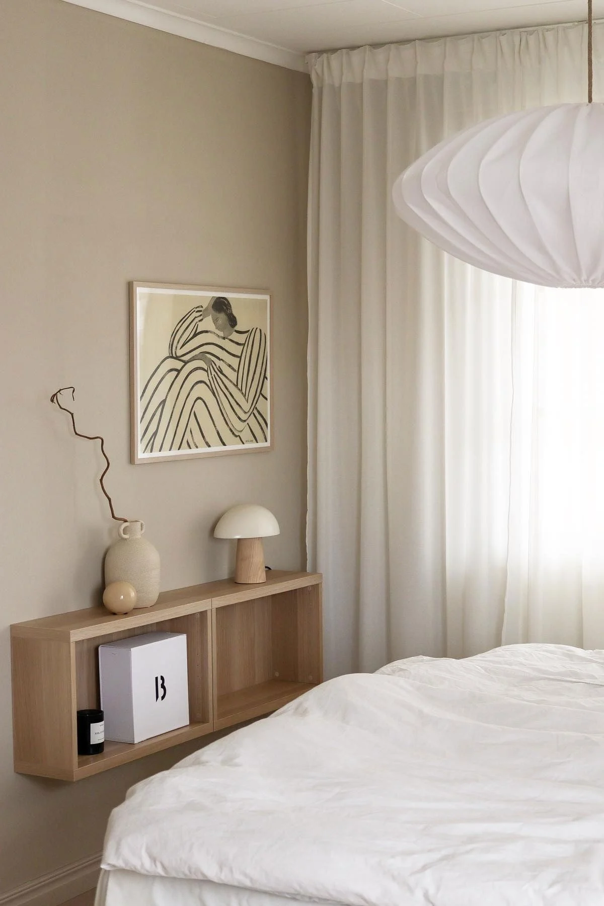 Minimalistisches Schlafzimmer mit weißer Leinenbettwäsche, schwebendem Holzregal mit Deko, abstrakter Kunst, transparenten Vorhängen und plissierter Hängelampe.