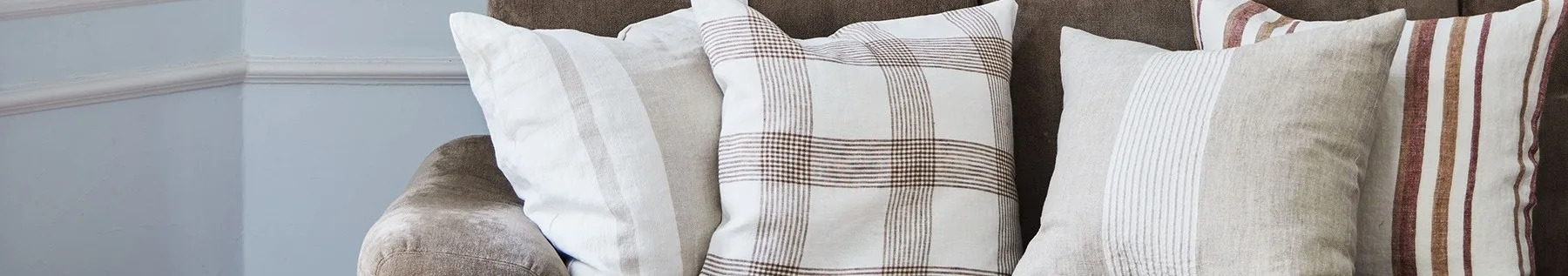 Nahaufnahme von drei Kissen auf einem braunen Sofa, mit weißen, beigefarbenen und karierten Mustern mit braunen und roten Streifen.