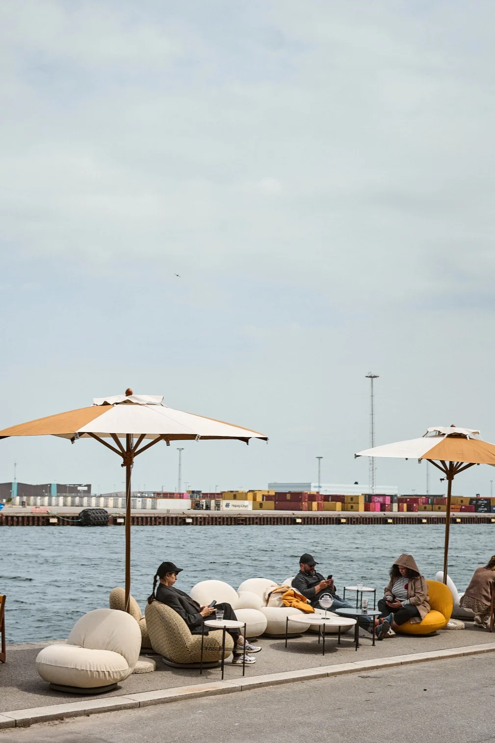 Menschen entspannen auf modernen Gartenmöbeln an einem Hafen mit Containern. Zwei Schirme spenden Schatten für die runden Stühle und Sofas.