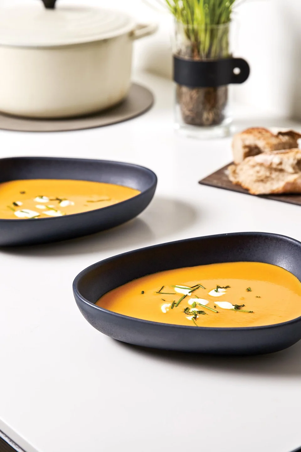 Zwei dunkle, ovale Schalen mit orangefarbener Suppe, Sahne und Schnittlauch auf einem weißen Tisch. Ein Topf, eine Pflanze und Brot sind im unscharfen Hintergrund.