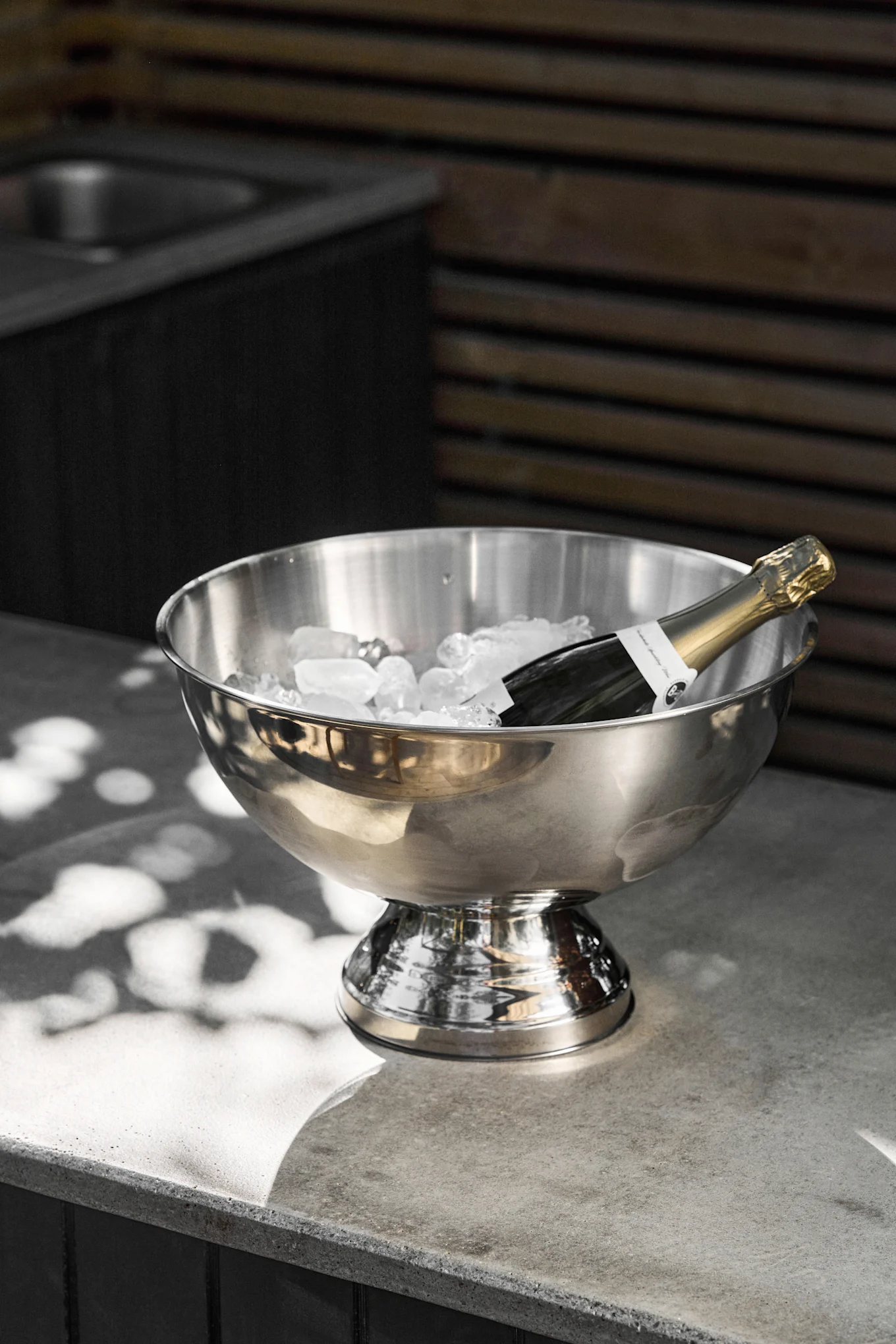 Eine Champagnerflasche liegt in einem Eiskübel aus Edelstahl, gefüllt mit Eiswürfeln, auf einer grauen Betontheke mit Sonnenflecken.