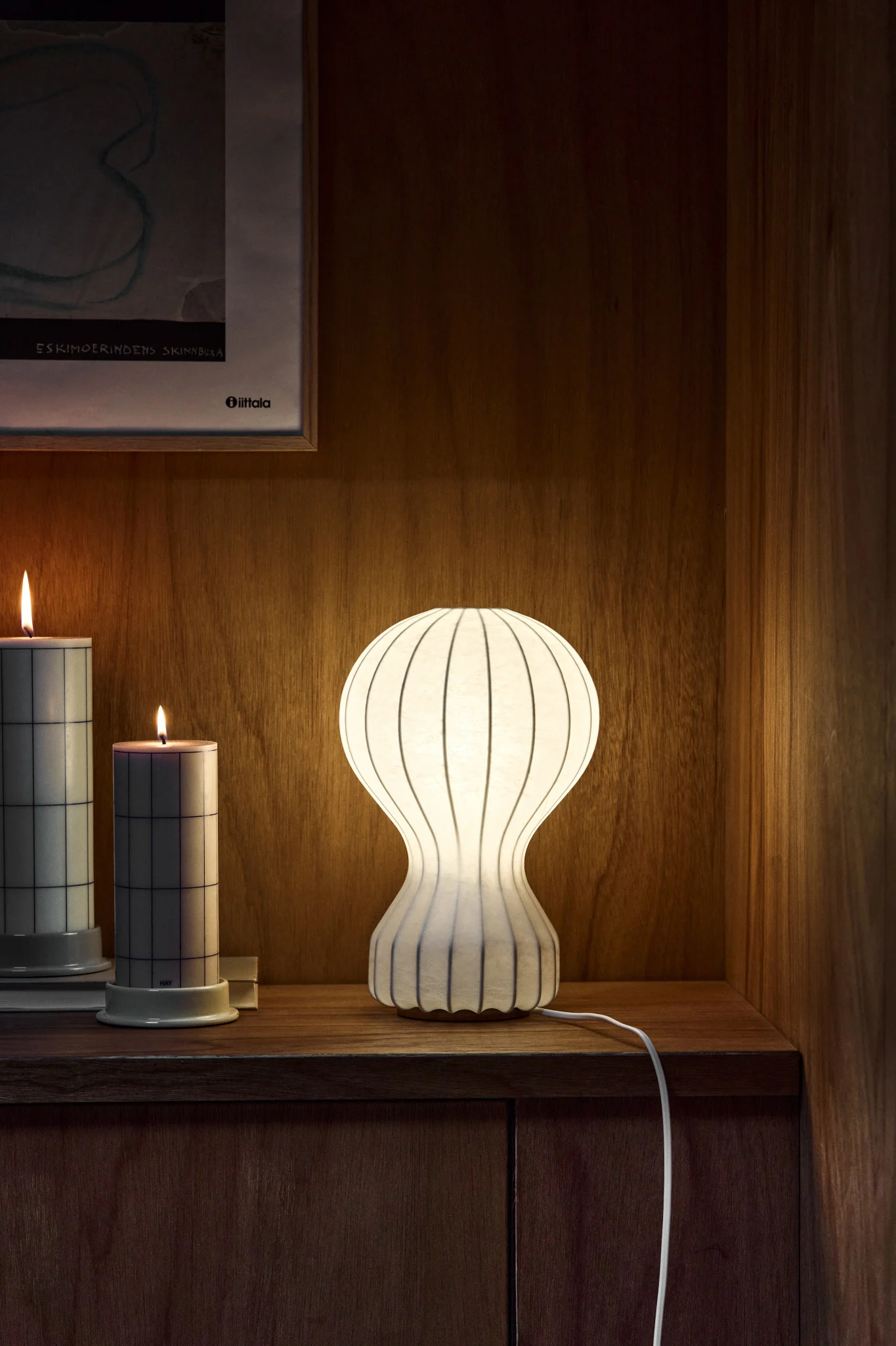 Beleuchtete weiße gerippte Tischlampe, zwei Kerzen mit Gittermuster und gerahmte Kunst auf Holzregal.