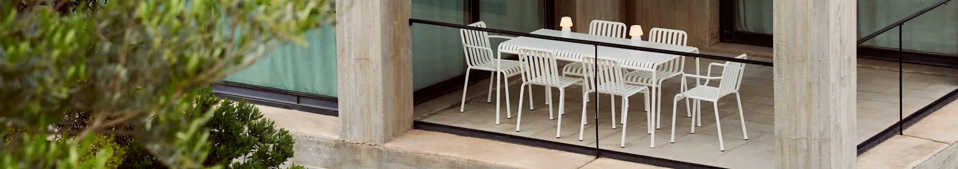 Eine Garten-Essgruppe mit einem weißen Lamellentisch und sechs passenden Stühlen, beleuchtet von drei tragbaren Lampen, auf einer Betonterrasse mit Pflanzen.