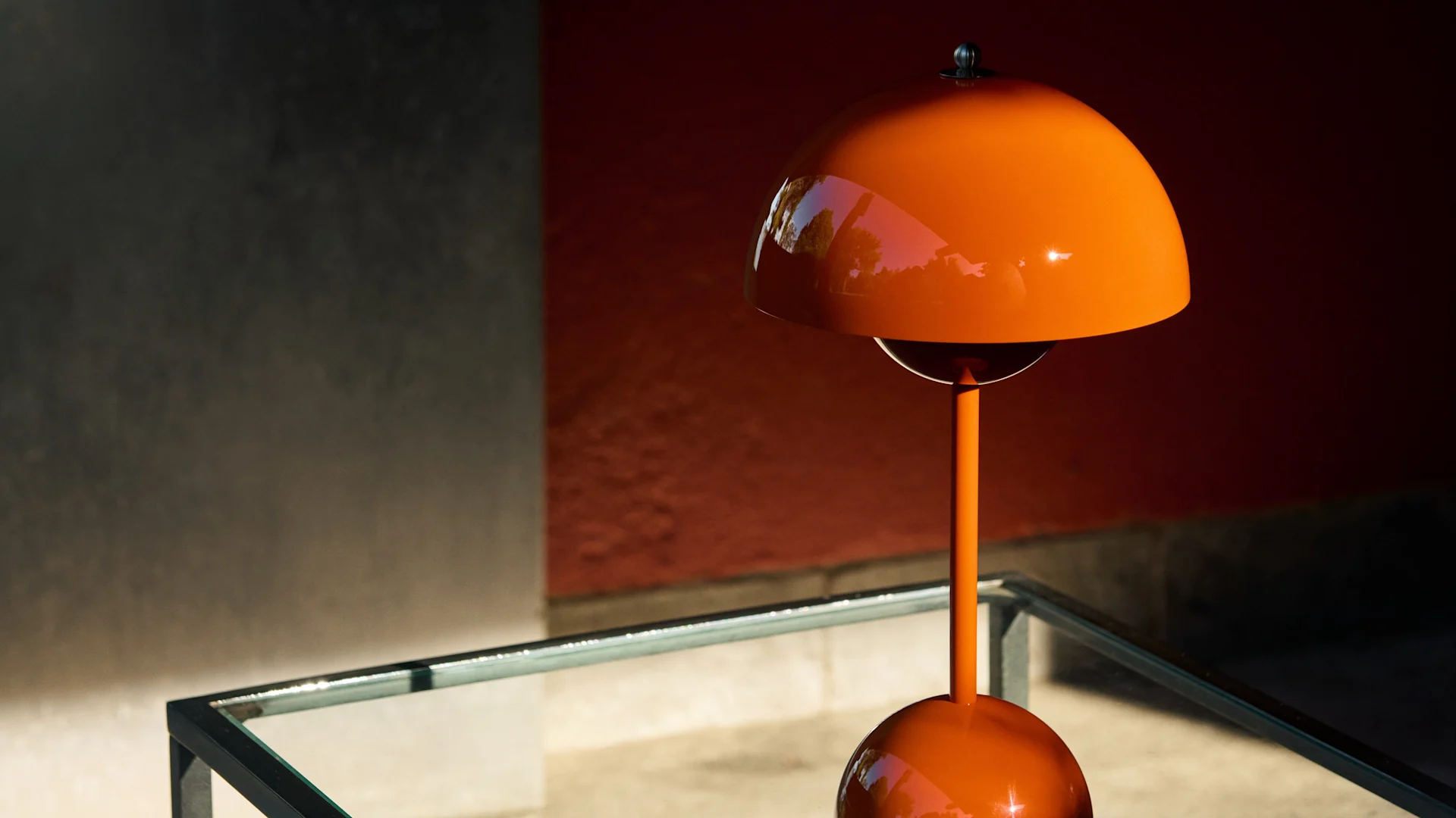 Glatte, orange pilzförmige Tischlampe auf einem Glastisch vor einer roten und grauen Wand, mit geflecktem Sonnenlicht.