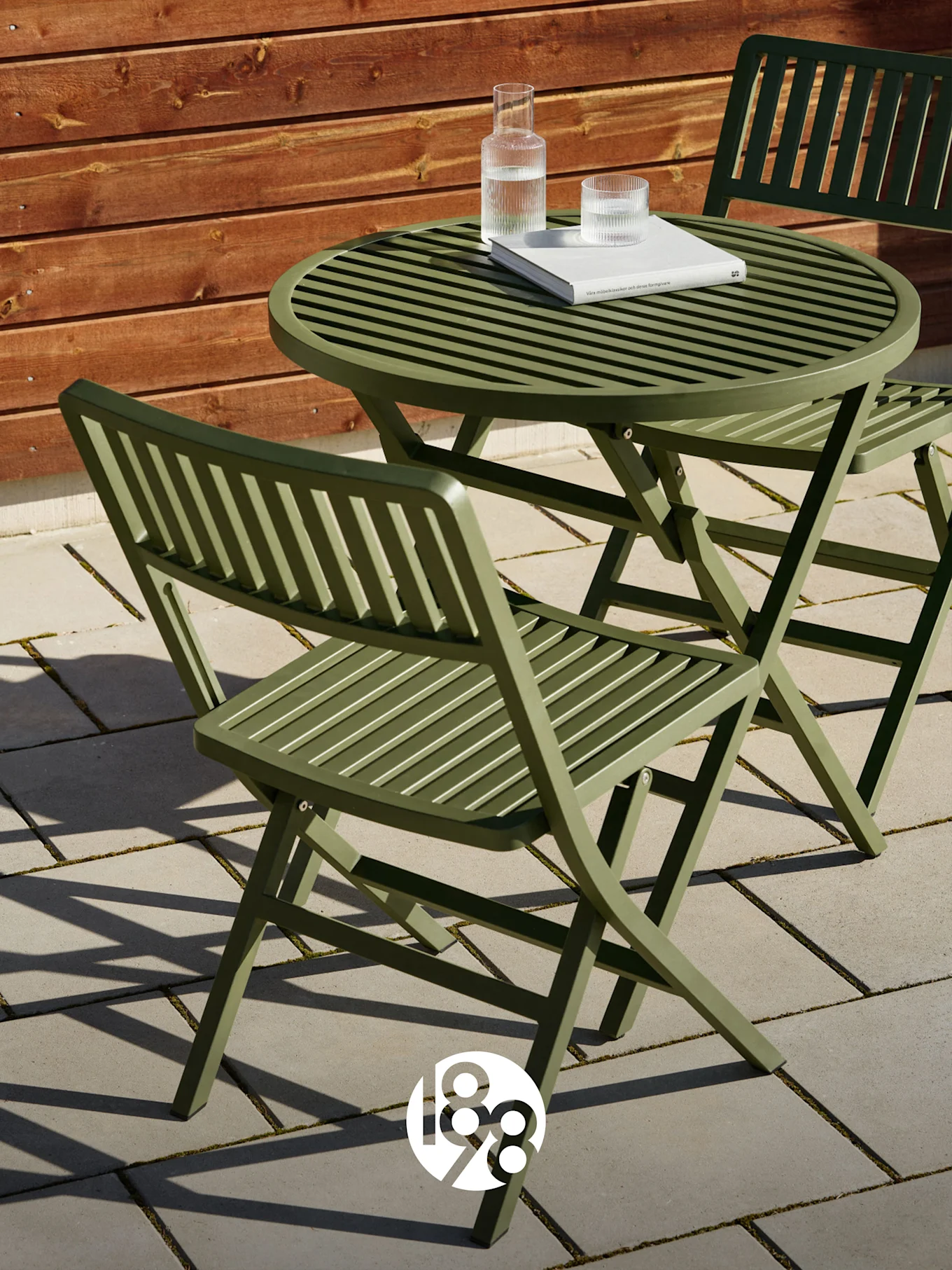 Zwei grüne Klappstühle mit Lamellen und ein passender Tisch auf einer Terrasse mit Buch und Wassergläsern.