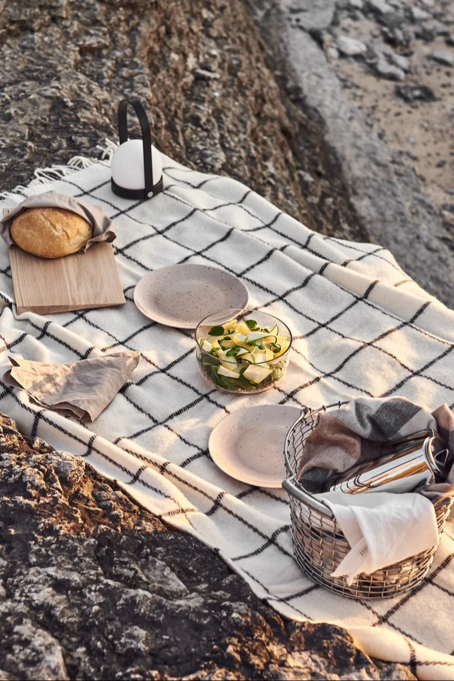 Picknick auf Felsen mit karierter Decke, Brot, Laterne, Salat, Tellern und Korb.