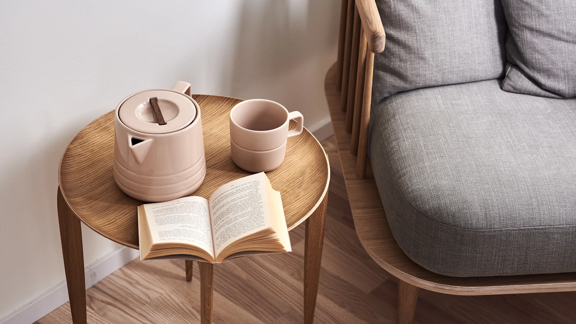 Ein hellrosa Teekannen-Set und ein offenes Buch auf einem hellen Holztisch neben einem grau gepolsterten Sessel mit Holzarmlehnen.