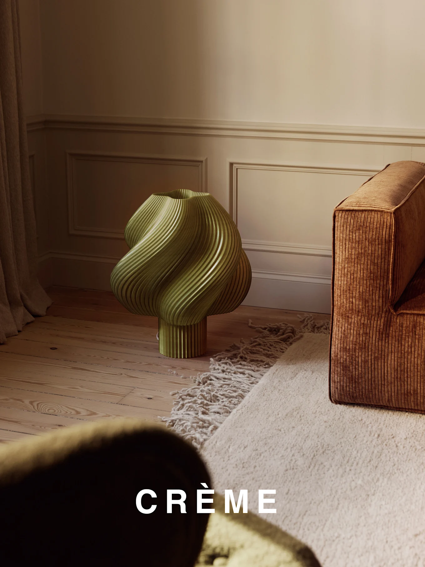 Eine hellgrüne, verdrehte gerippte Lampe auf einem Holzboden, neben einem braunen gerippten Sofa und einem beigen Zottelteppich.