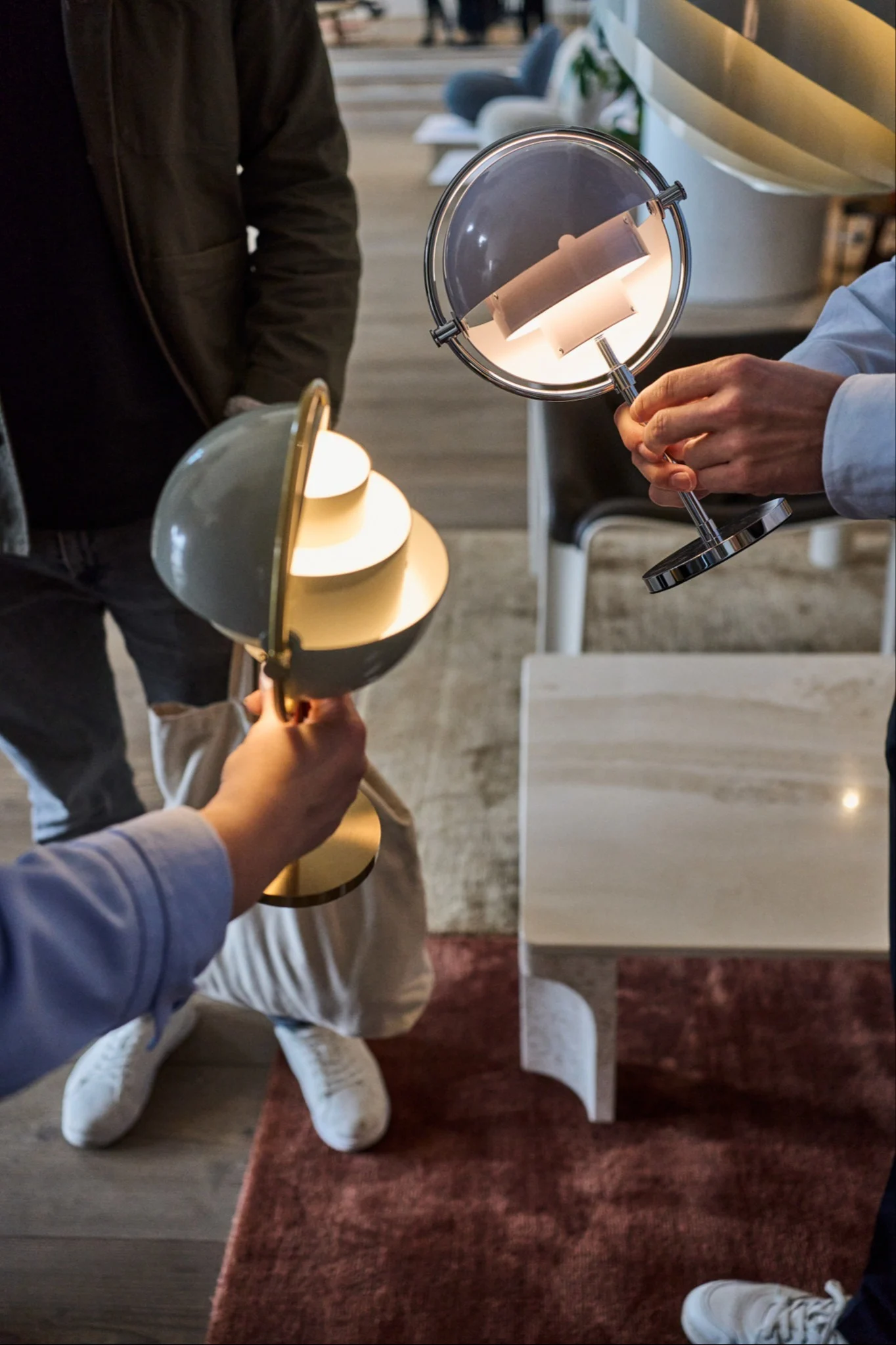 Zwei Personen halten beleuchtete moderne Schreibtischlampen, eine mit grauem kugelförmigem Schirm und goldenem Sockel, die andere mit rundem grauem Schirm und silbernem Rahmen.