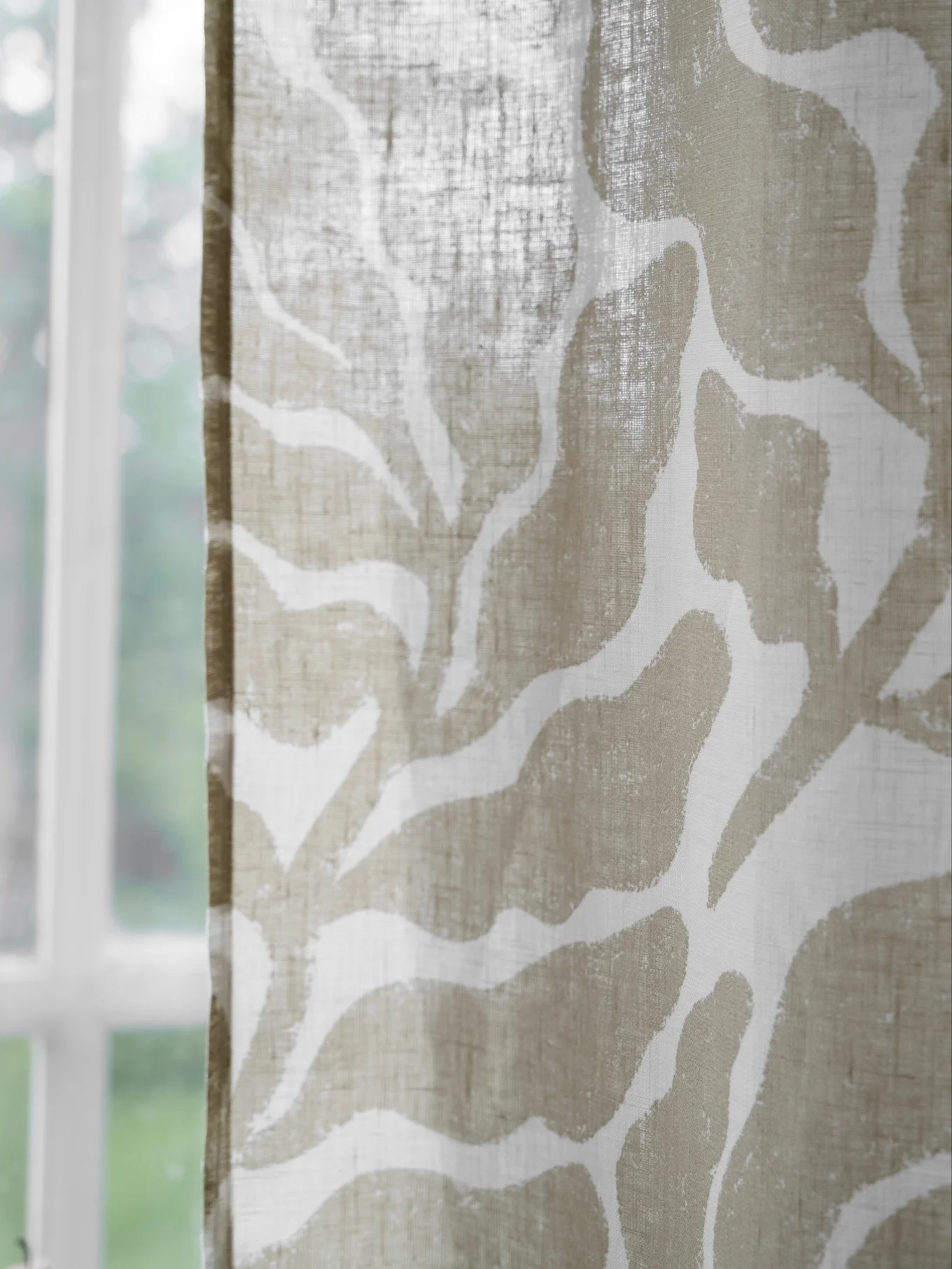 Nahaufnahme eines transparenten Fenstervorhangs mit beigem, abstraktem Muster, im Hintergrund ein verschwommenes Fenster und Grün.