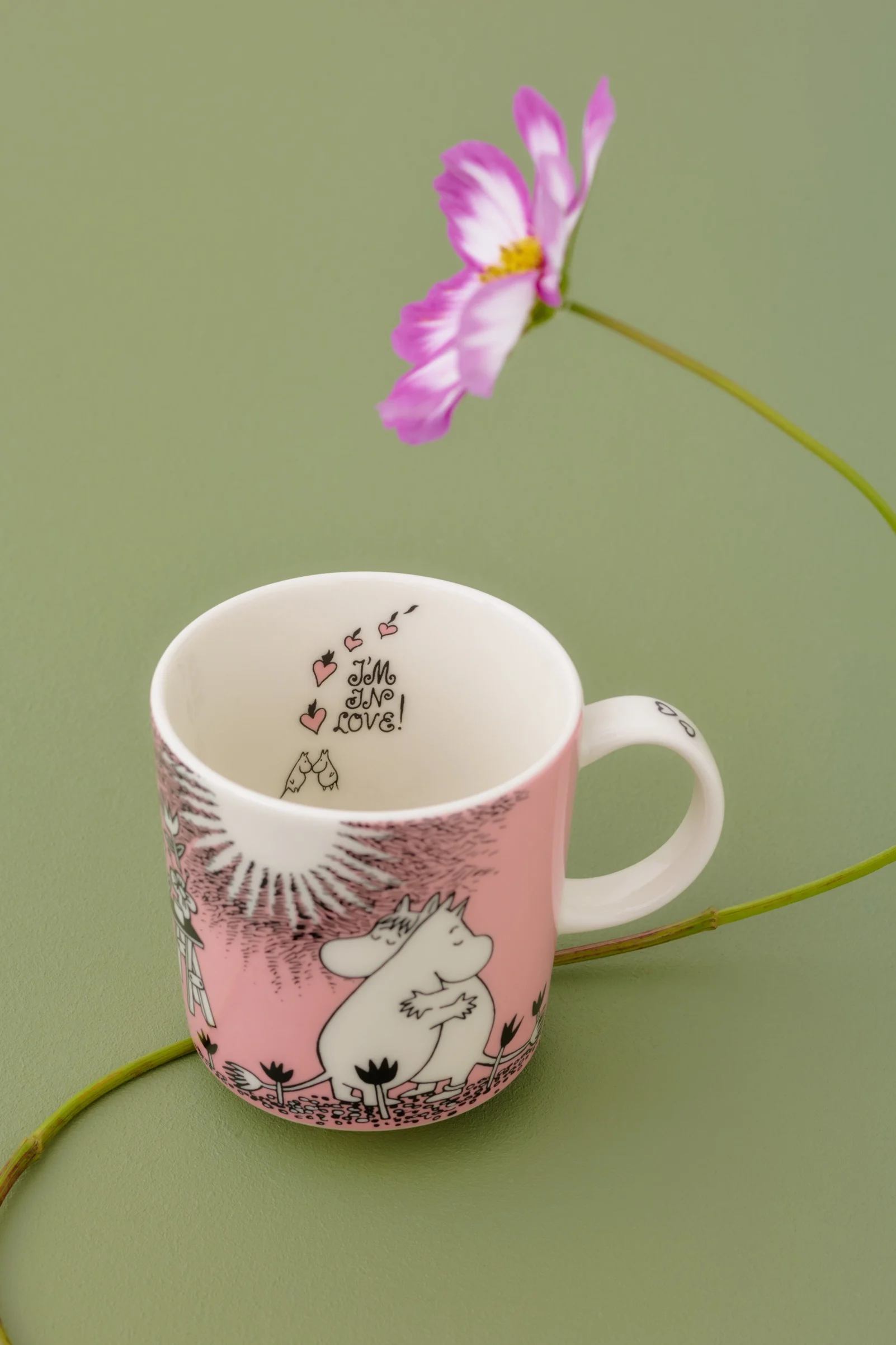 Rosa-weiße Moomin-Tasse mit Moomintroll und Snorkfräulein, "I'M IN LOVE!" Text innen, auf grünem Hintergrund mit Kosmosblume.