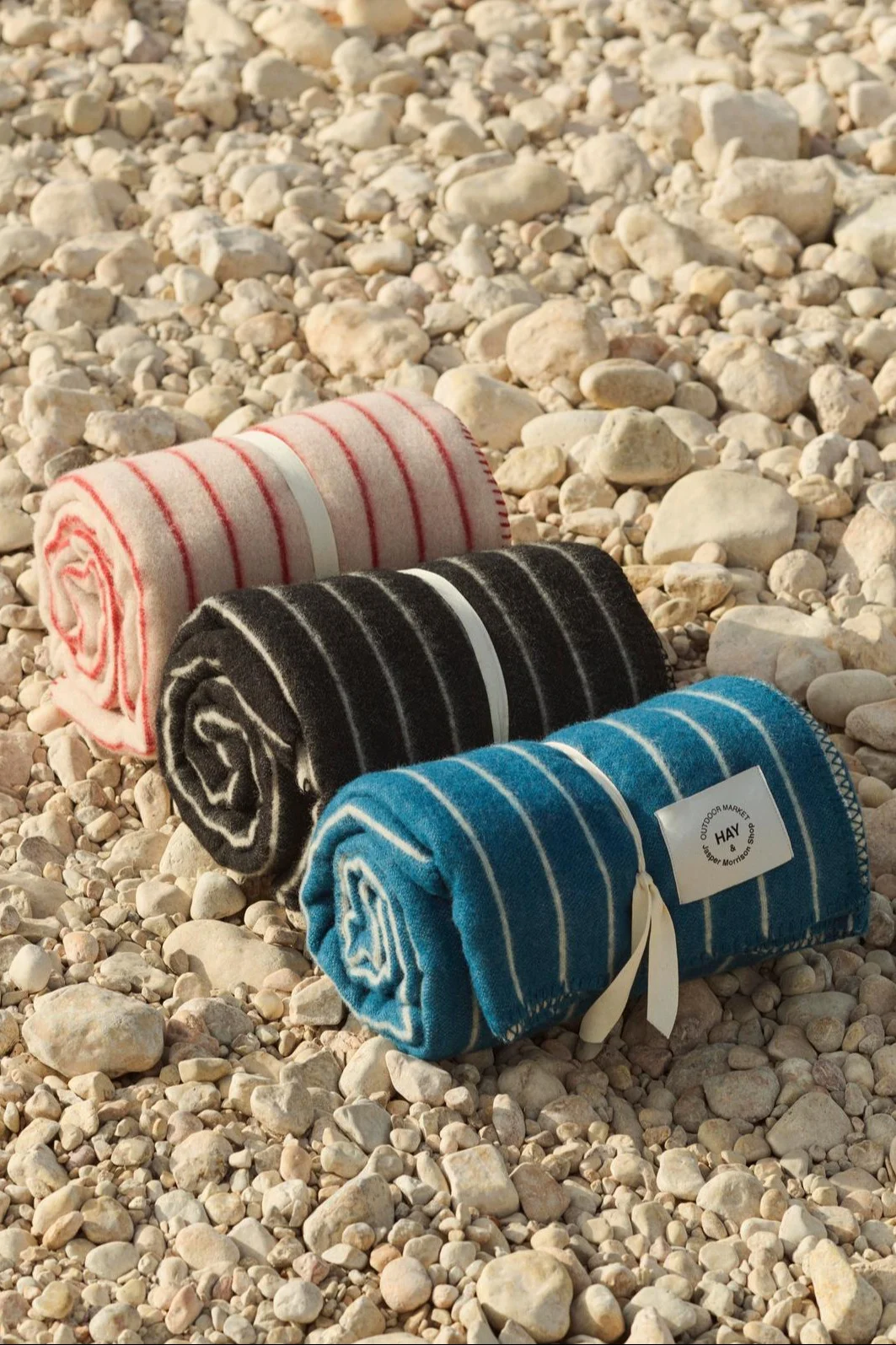 Drei gerollte gestreifte Decken in Pink, Schwarz und Blau, mit Bändern auf einem Kiesstrand.