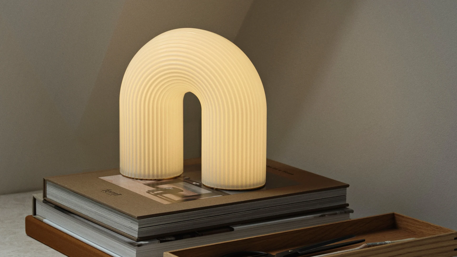 Warm leuchtende, bogenförmige Tischlampe mit gerippter Textur auf einem Stapel brauner Bücher, mit einer Holzkassette daneben.
